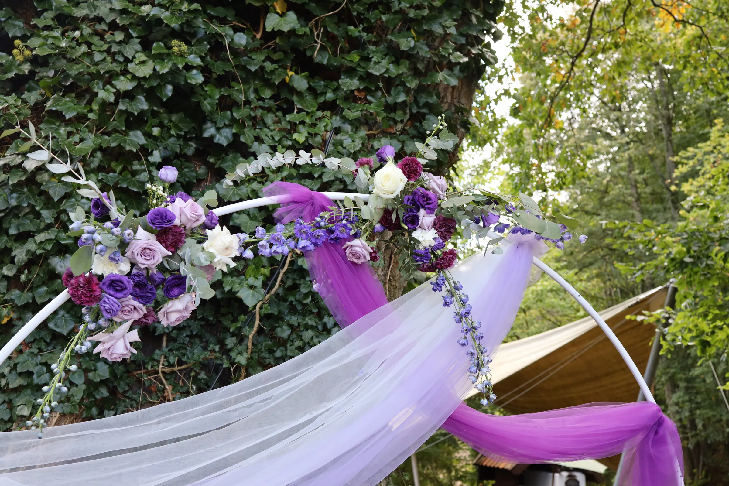 Composition of roses, eustoma and delphiniums on a white rim with lilac and purple tulle, old trees in the background