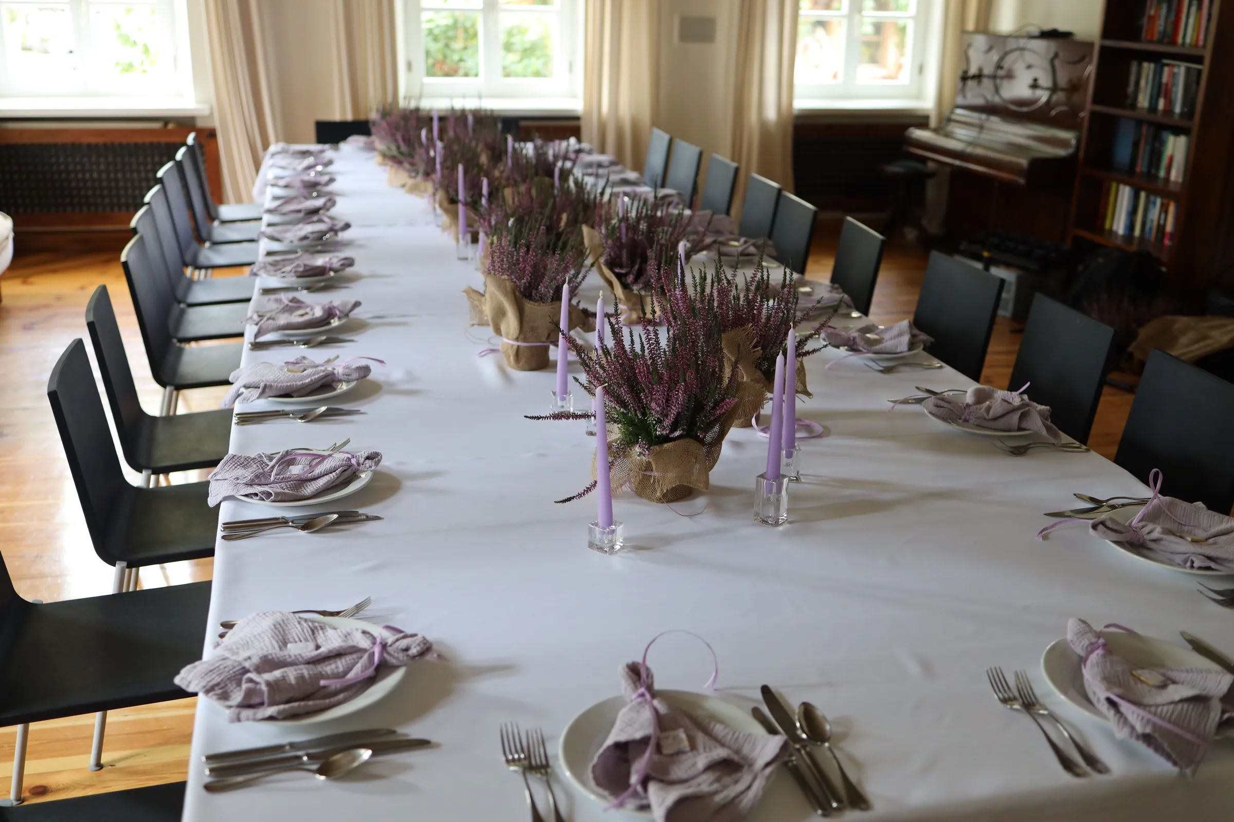 A long table with a white tablecloth in a room with windows and a piano, in the middle a row of heather in jute and lilac candles