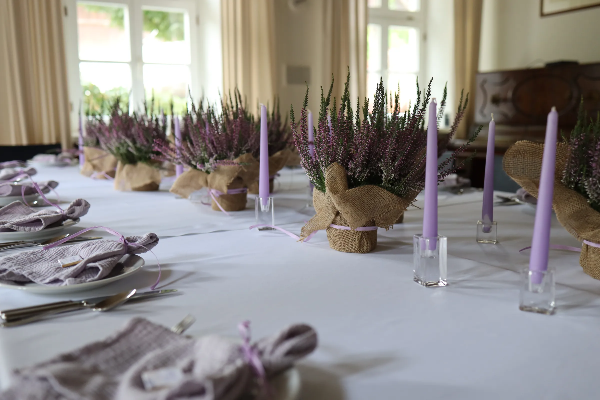 Heathers in jute casings and lilac candles in glass candlesticks along a long white table, with napkins tied with a lilac ribbon at the place settings.