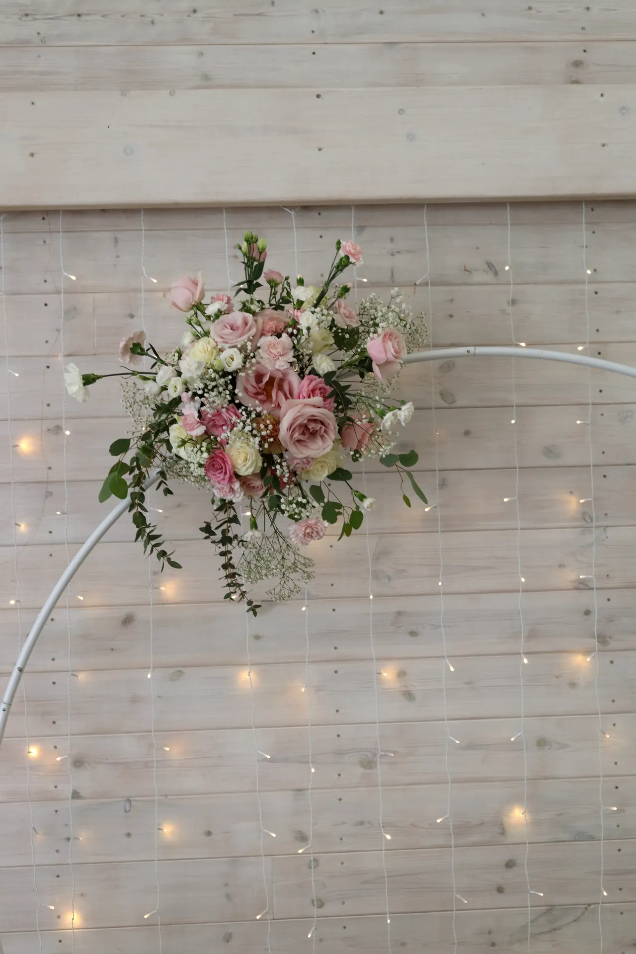 A white rim with a cluster of pink and cream flowers and greenery against the background of light boards and warm lamps