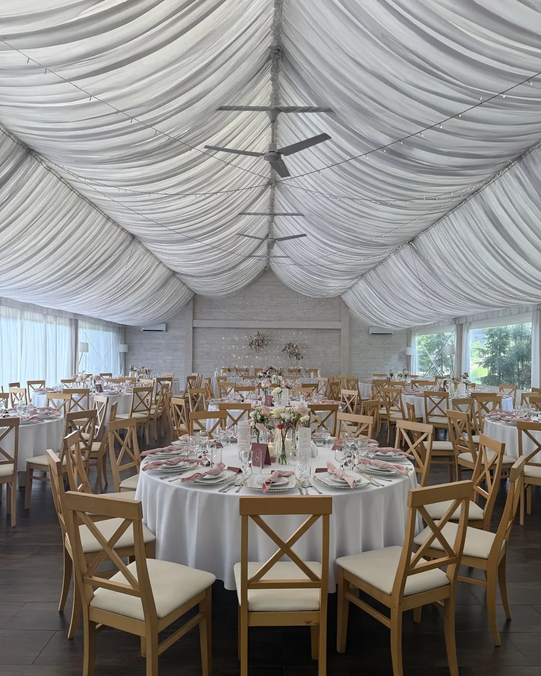 A bright room under a white draped ceiling, round white tables, wooden chairs and pink napkins