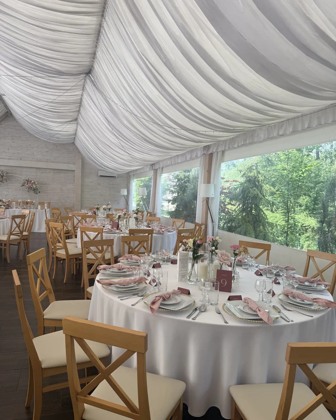Part of the room with large windows overlooking the garden, round tables with pink napkins and flowers in vases