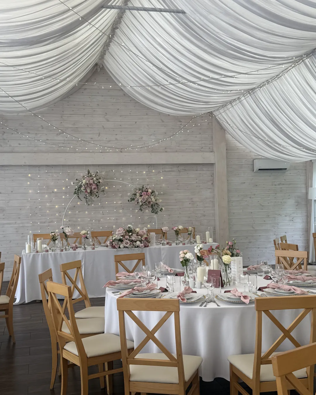 A view of the bride and groom's table with two flower hoops and a wall of lights, with round tables of guests in front of it