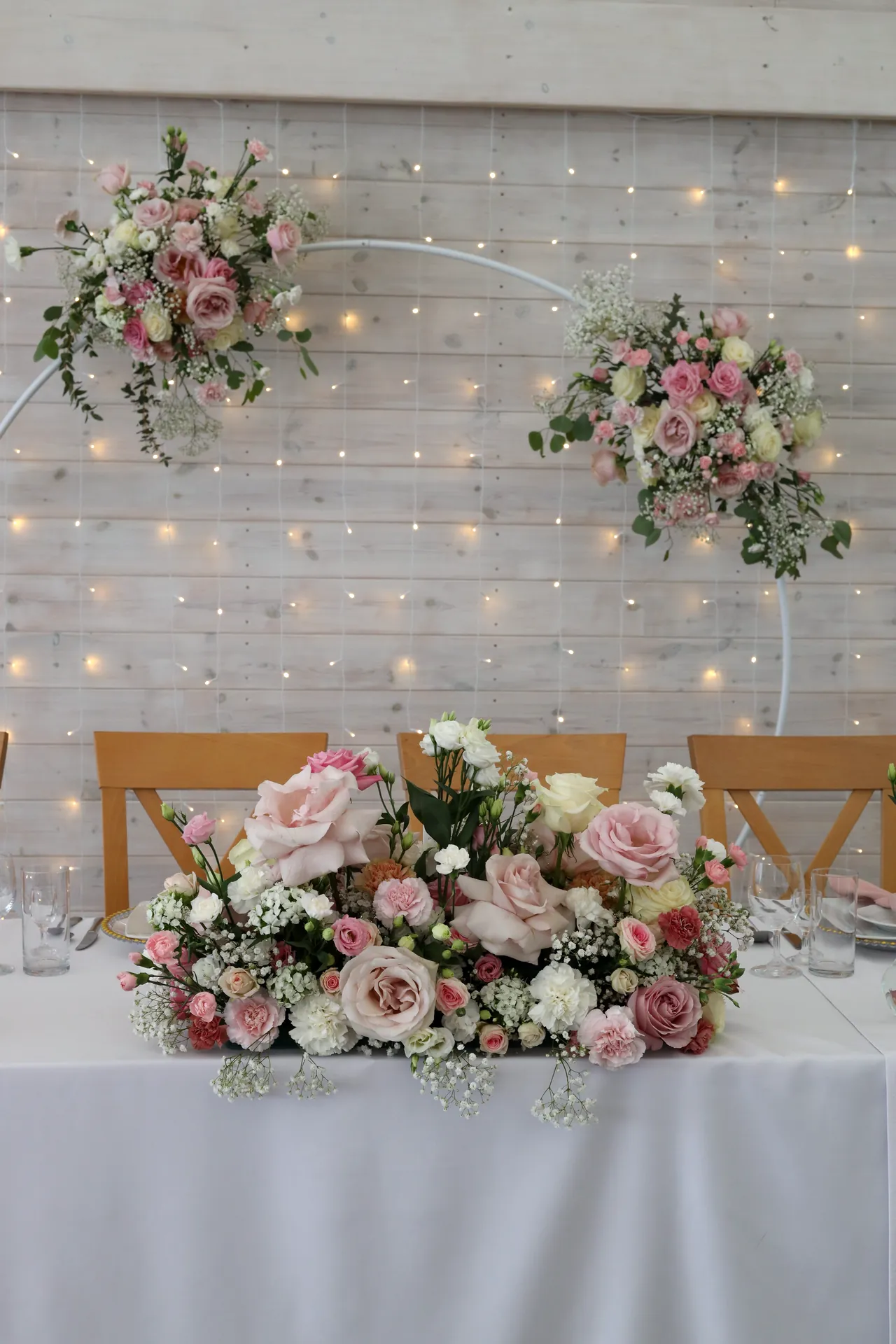 The bride and groom's table with a long composition of roses and gypsophila, behind it two white rings with flowers and warm lamps