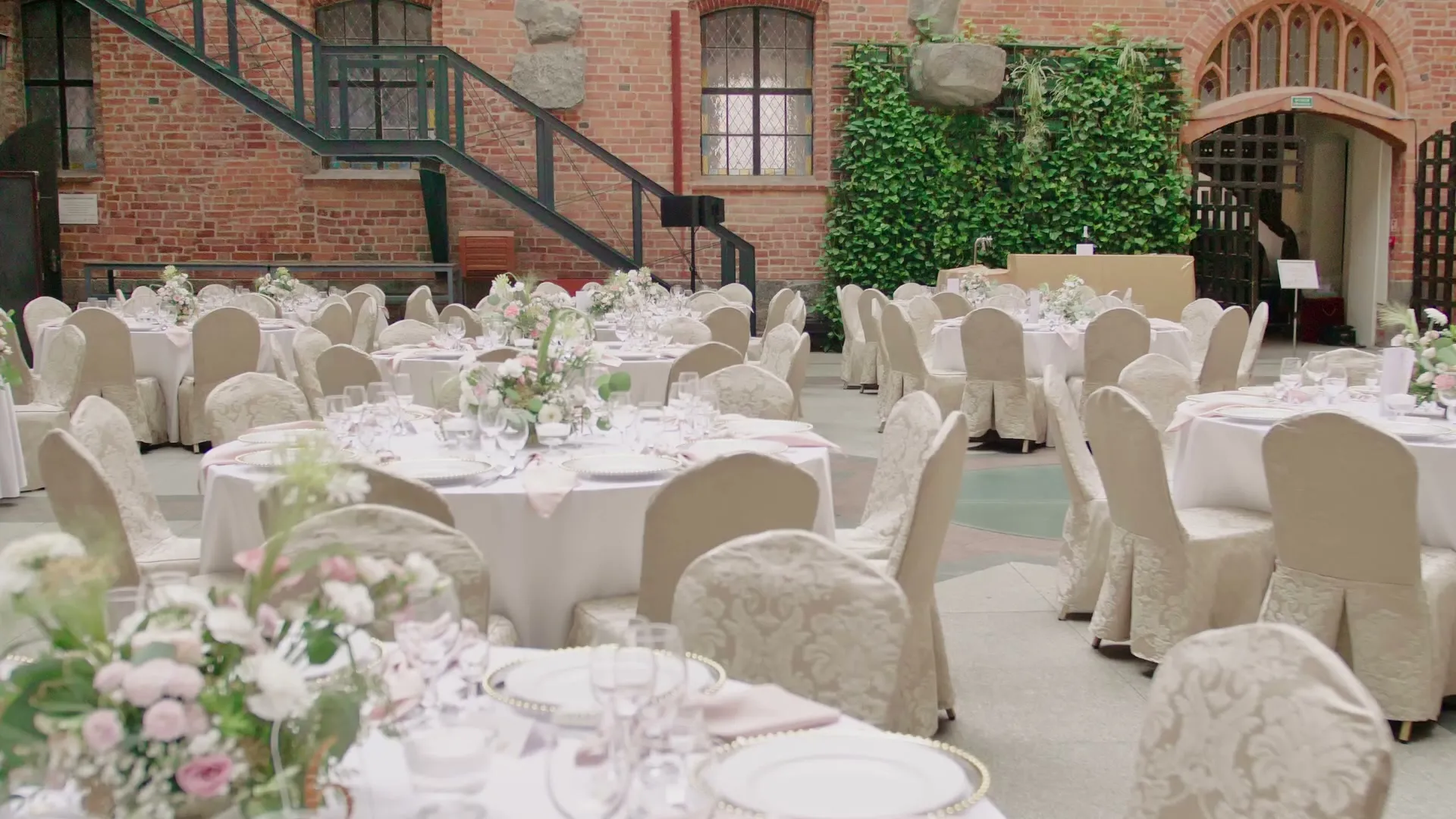 Round tables with low arrangements in the castle courtyard, a brick wall covered with ivy and wrought iron stairs in the background