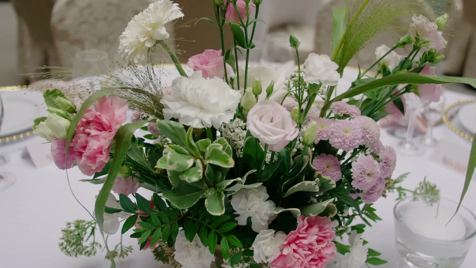 A low composition of pink carnations, eustomas and pom-pom chrysanthemums with ornamental grass, a floating candle in glass next to it