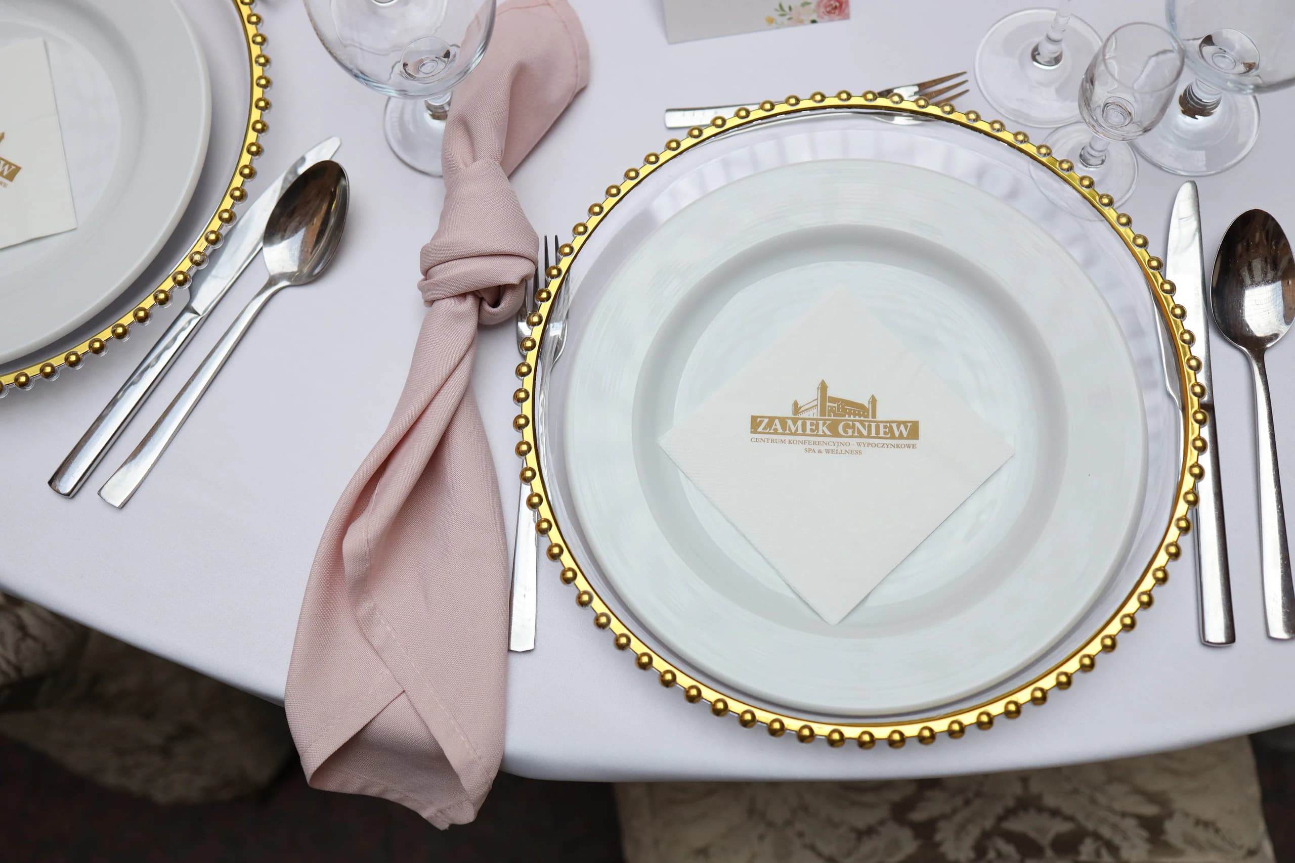Top setting: glass plate with gold pearl rim, white plate with a paper napkin with a castle print, pink knotted napkin and cutlery