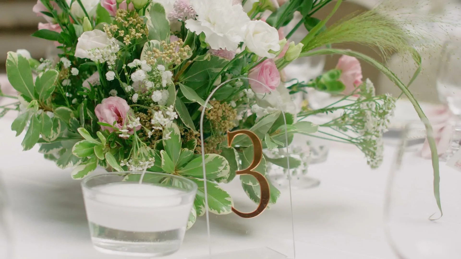 An acrylic plaque with a golden number 3 next to a low composition with pink eustomas and gypsophila, a floating candle in glass in front of it