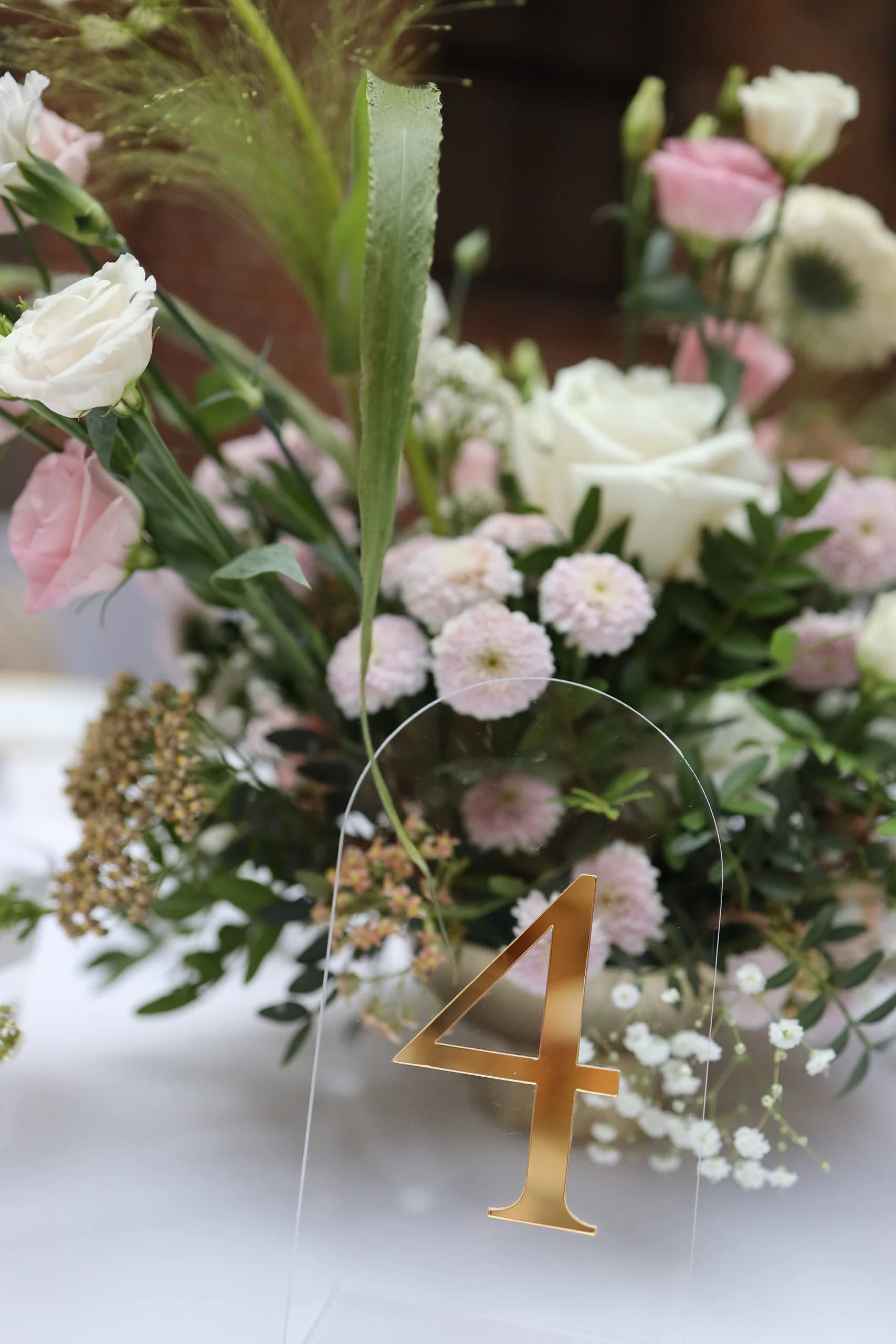 A transparent acrylic plaque in the shape of an arch with a gold number 4, behind it pink chrysanthemums, a white rose, eustomas and gypsophila