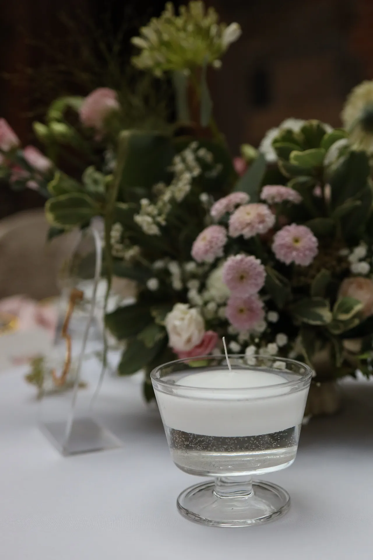 A floating candle in a glass cup on a leg, behind it a low composition of pink pompom chrysanthemums, gypsophila and greenery