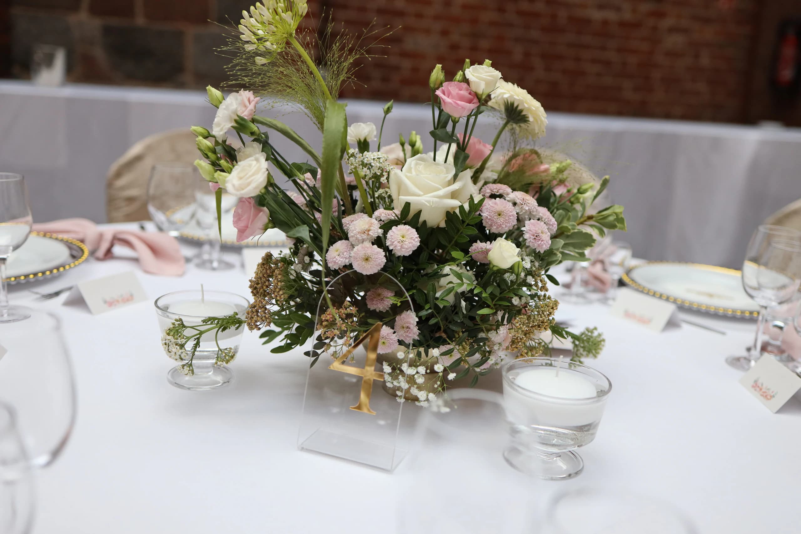 A low flower arrangement in a gold vessel, in front of it an acrylic plaque with a gold number 4, floating candles in glass cups on the sides