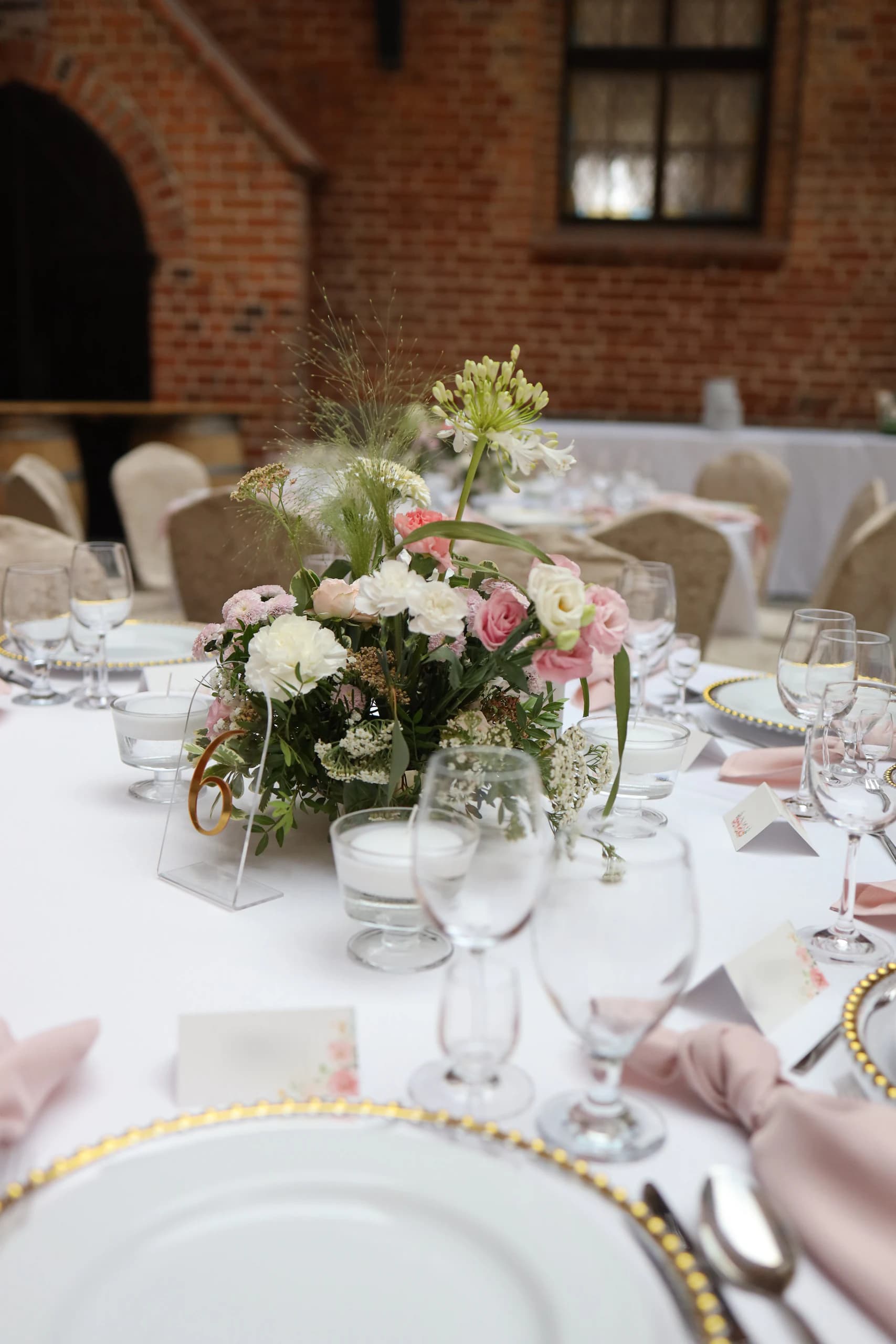 A round table with a low flower arrangement, an acrylic number 6 and floating candles in glass, behind it a pointed archway and a brick wall with a window