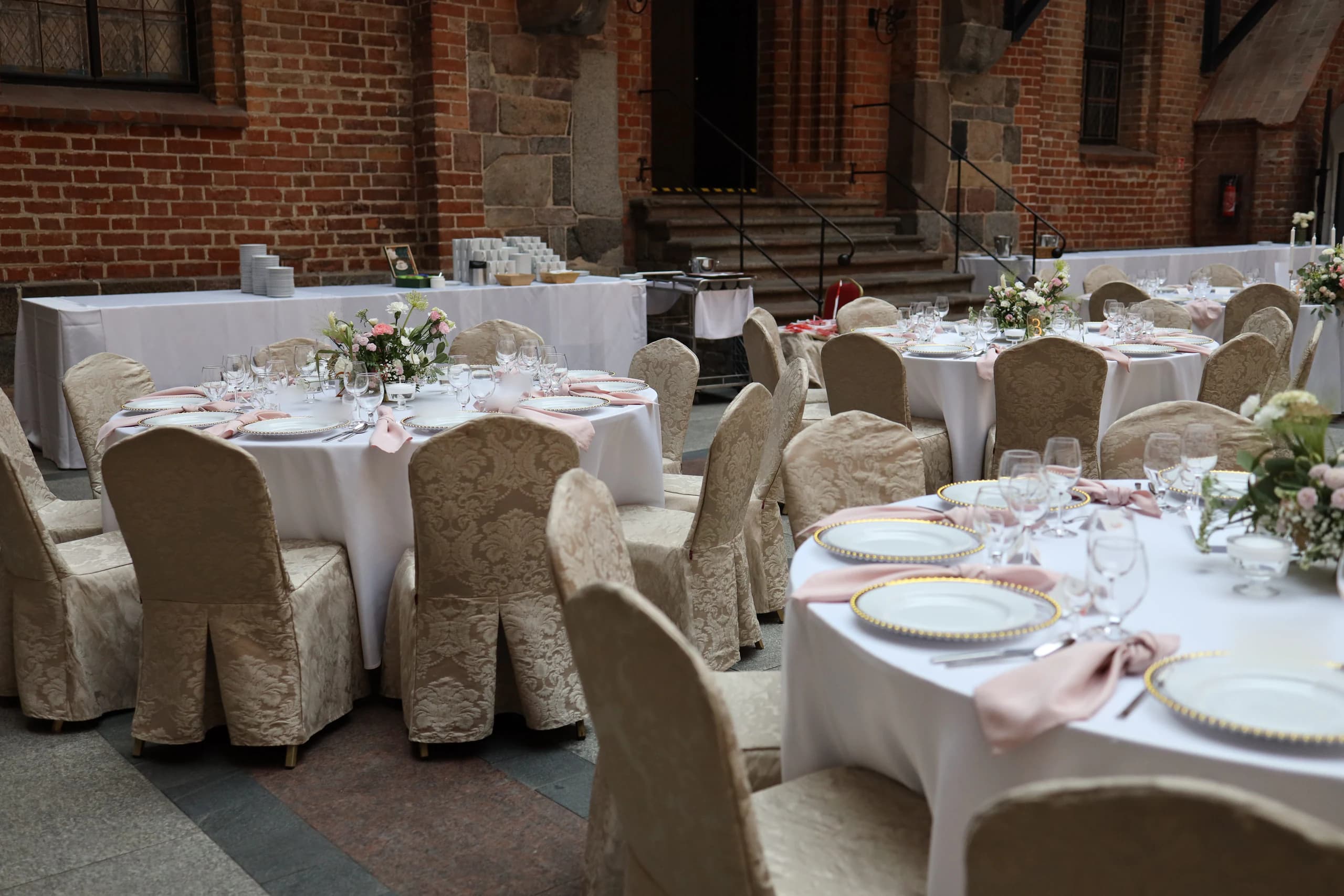 Round tables with white tablecloths and chairs with beige, jacquard covers in the castle courtyard, brick walls and stone stairs in the background