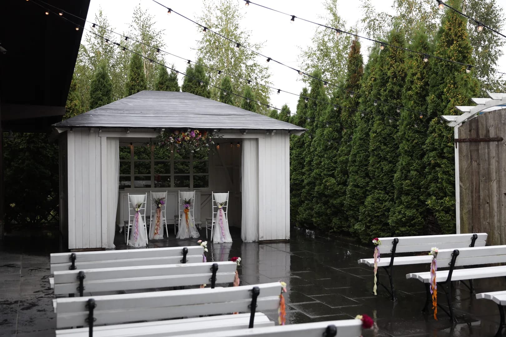 White gazebo with a flower arrangement under the eaves, four decorated chairs and white benches with bouquets on a wet square