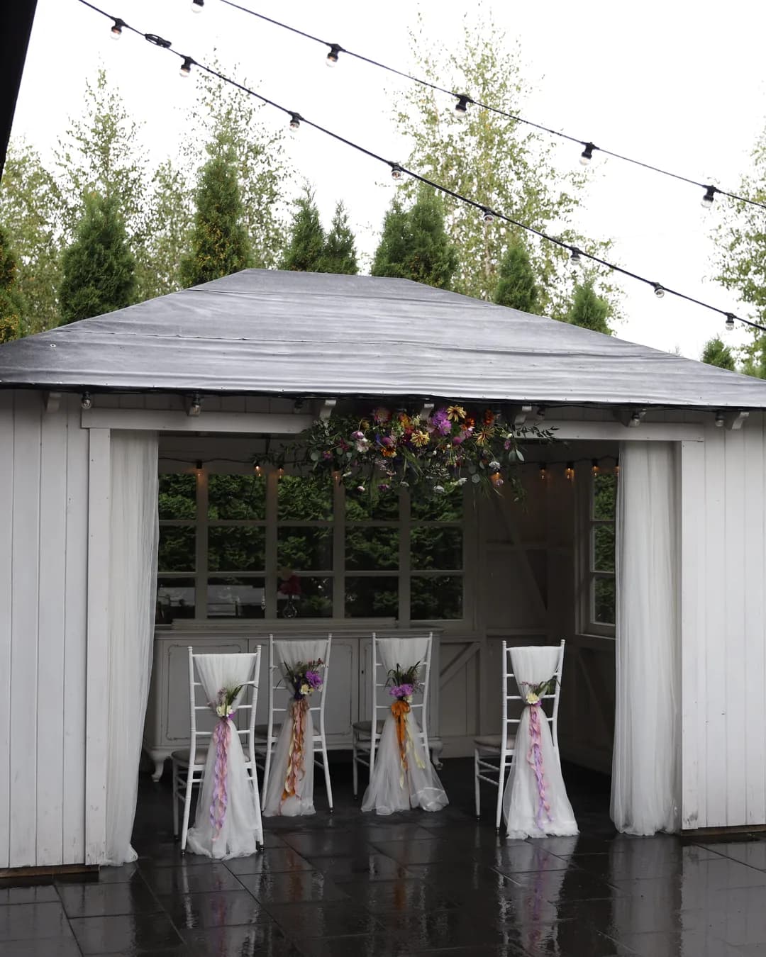 Gazebo with a hanging composition of wild flowers and four chairs with tulle, bouquets and long ribbons
