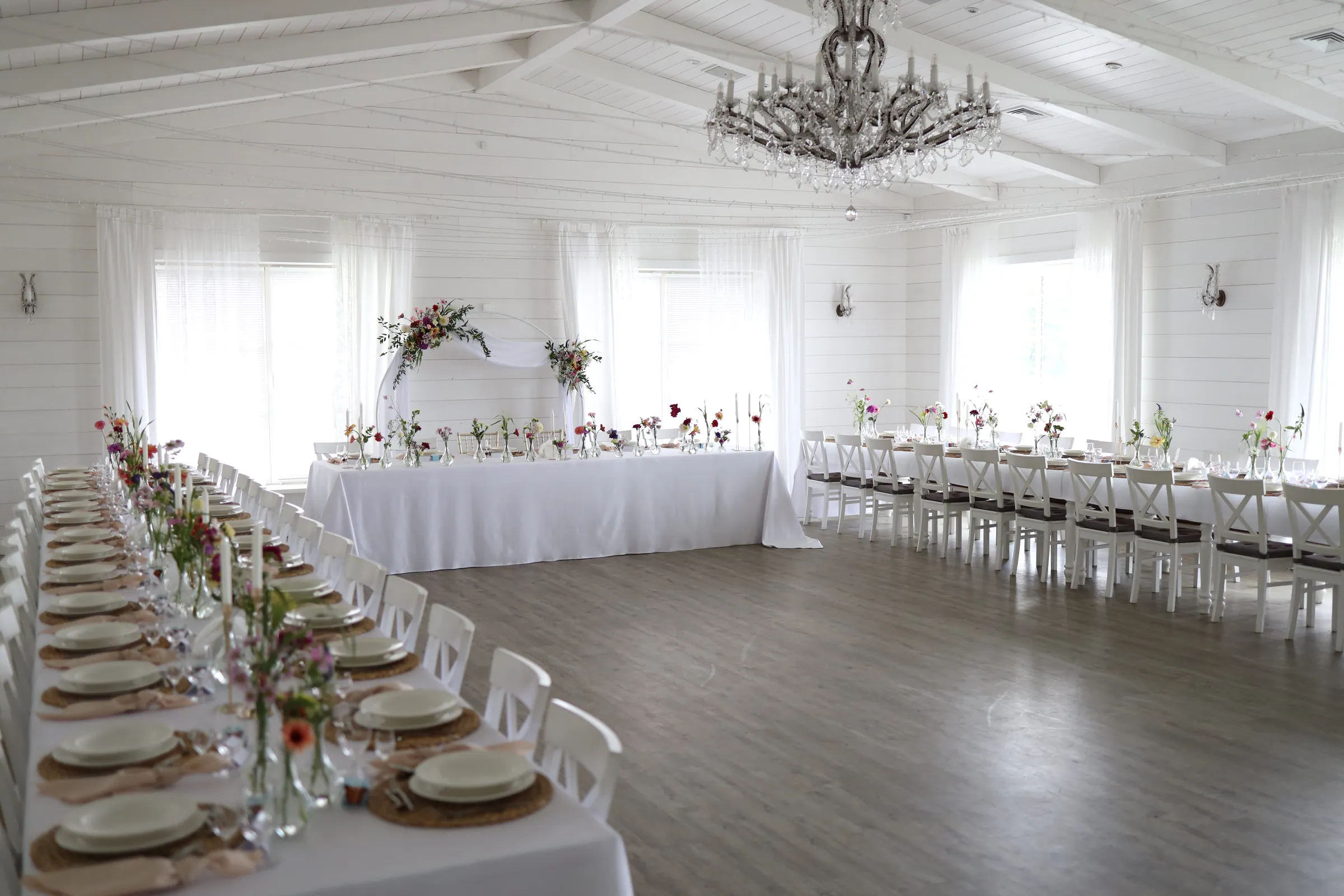A bright room with white board walls and a beamed ceiling, long guest tables with vases and white chairs