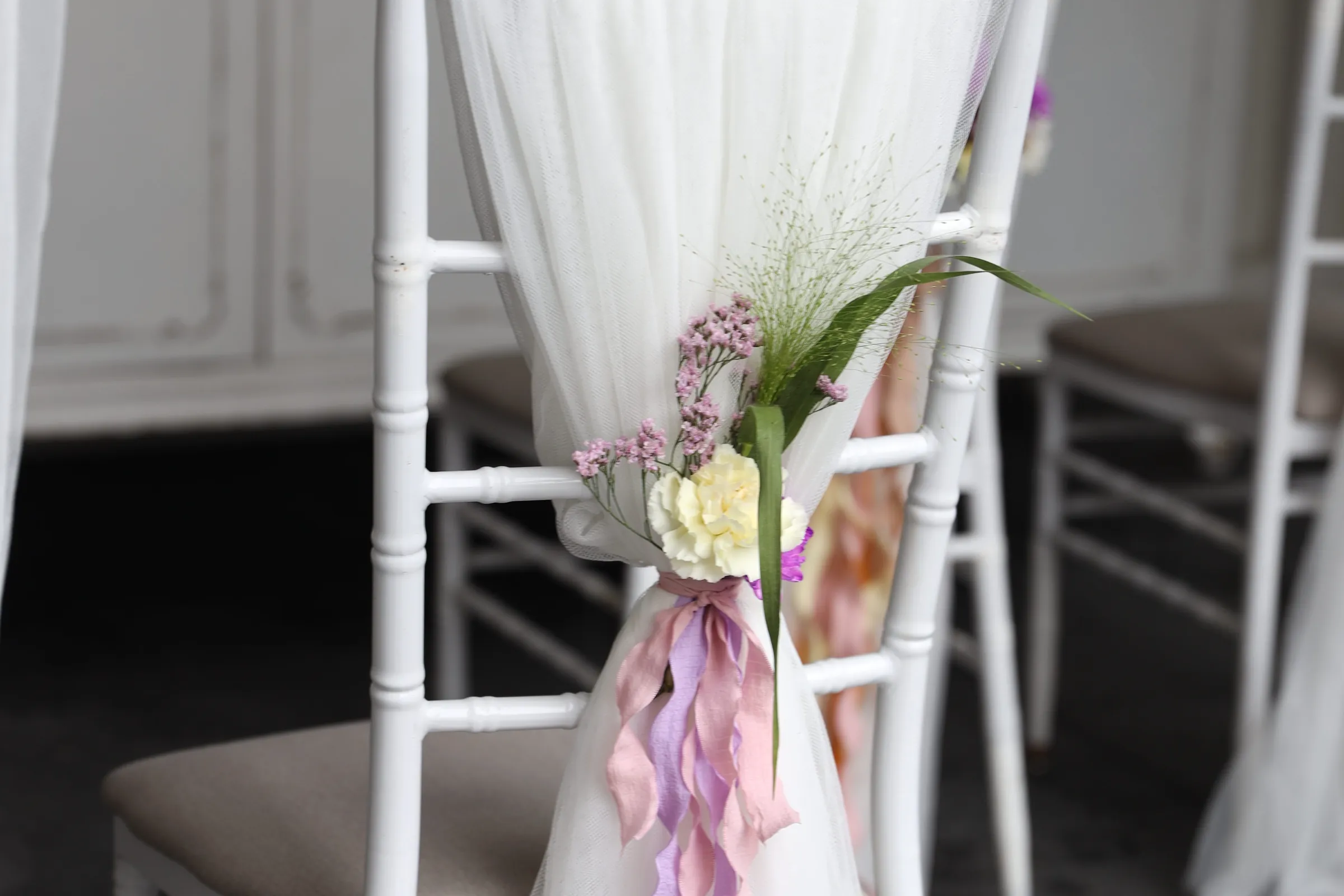 Chair backrest in white tulle with a bouquet of cream carnations, tiny pink flowers and grasses, and wavy ribbons