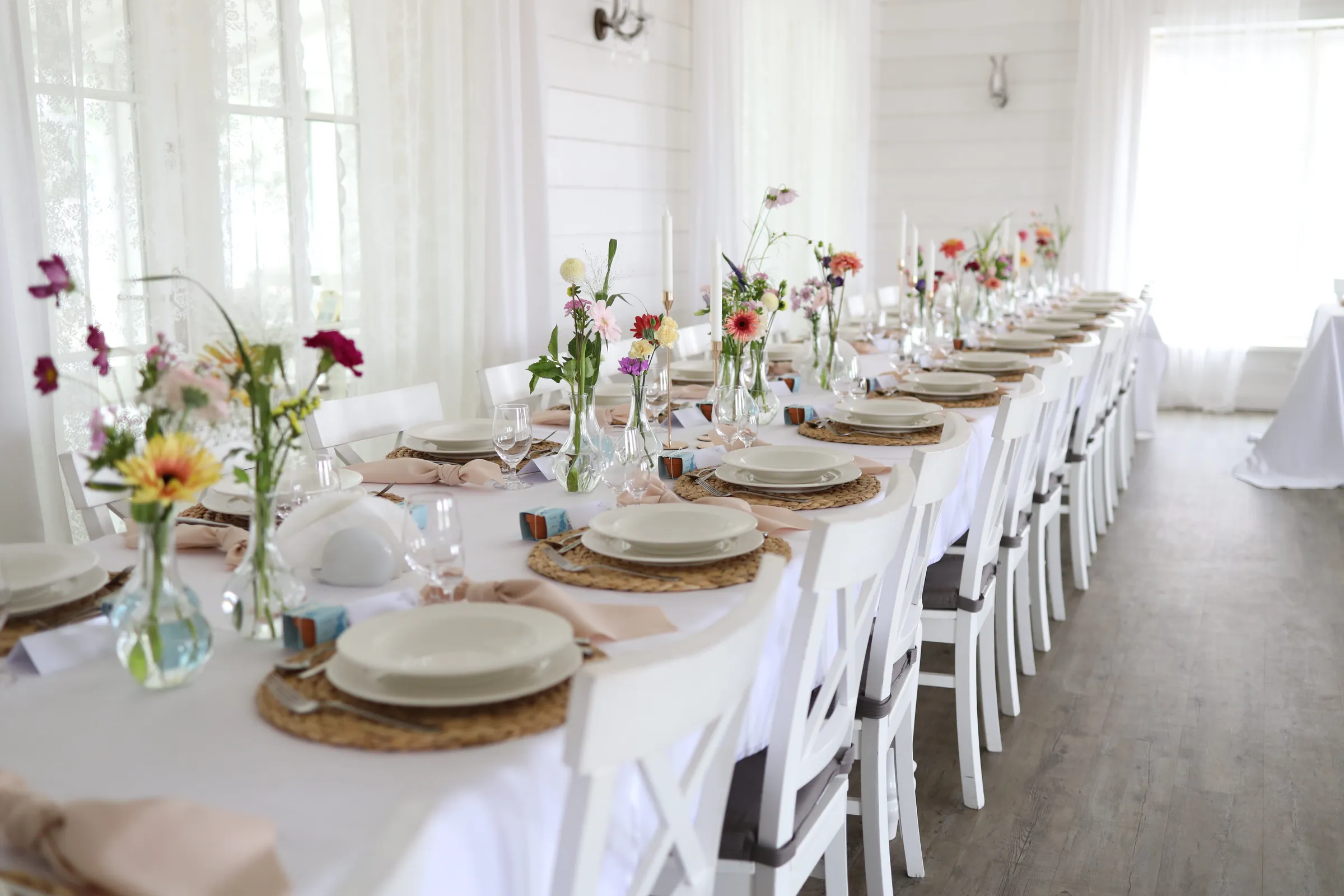 A long guest table with a row of glass vases with wild flowers, woven table plates and peach napkins