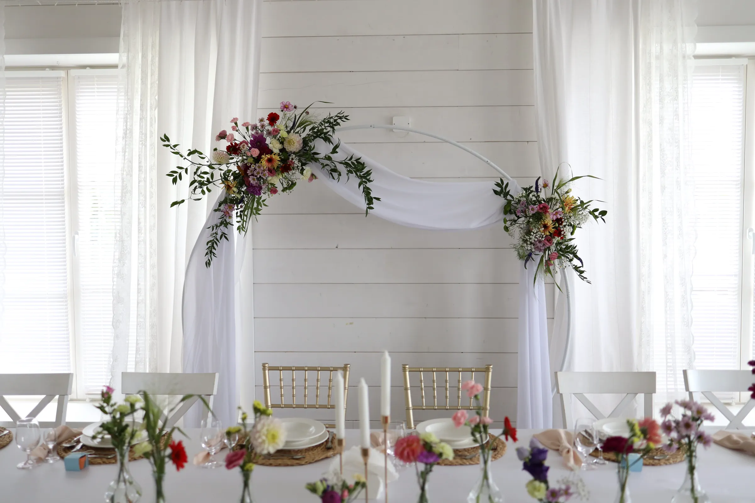 A white circle with tulle in front of a wall of white boards, a large wildflower arrangement on the left and a smaller one on the right