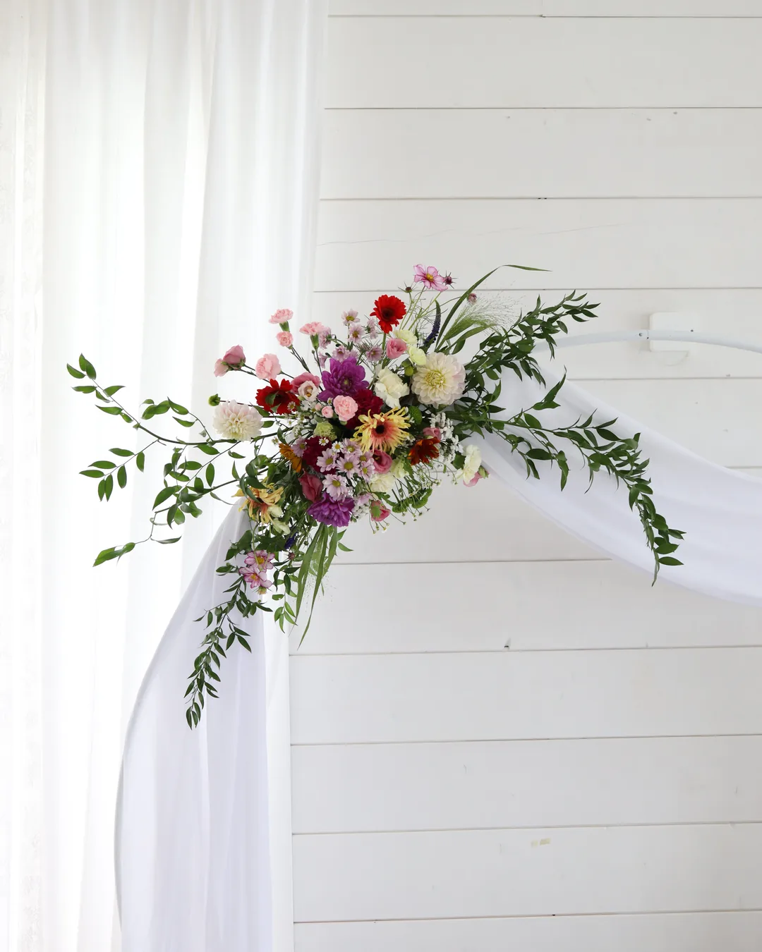 Composition of dahlias, gerberas, carnations, chrysanthemums and cosmoses with long greenery on a white circle with draped tulle