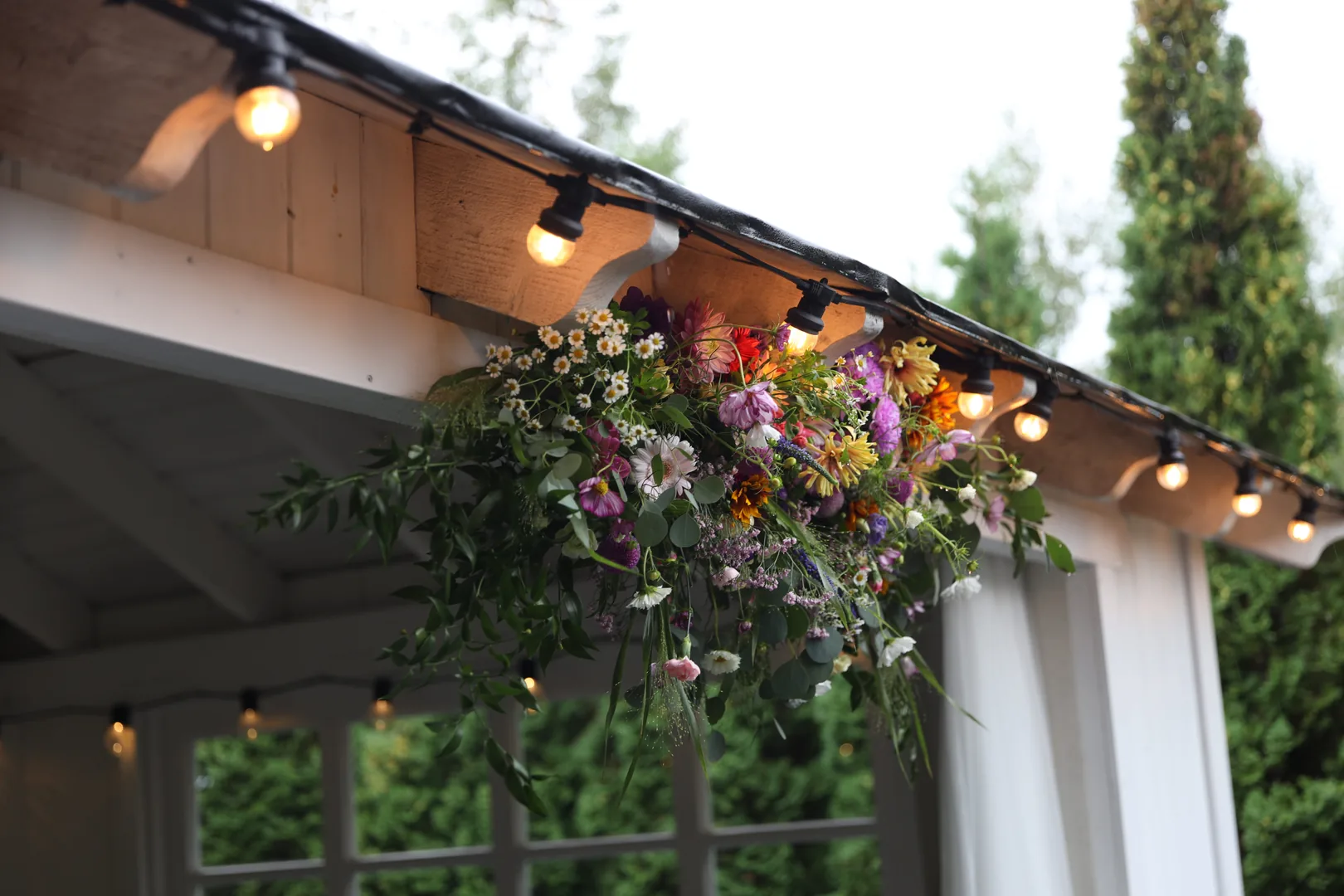 A composition of dahlias, cosmoses, gerberas and small white flowers with greenery under the eaves of the gazebo, with warmly glowing light bulbs next to them