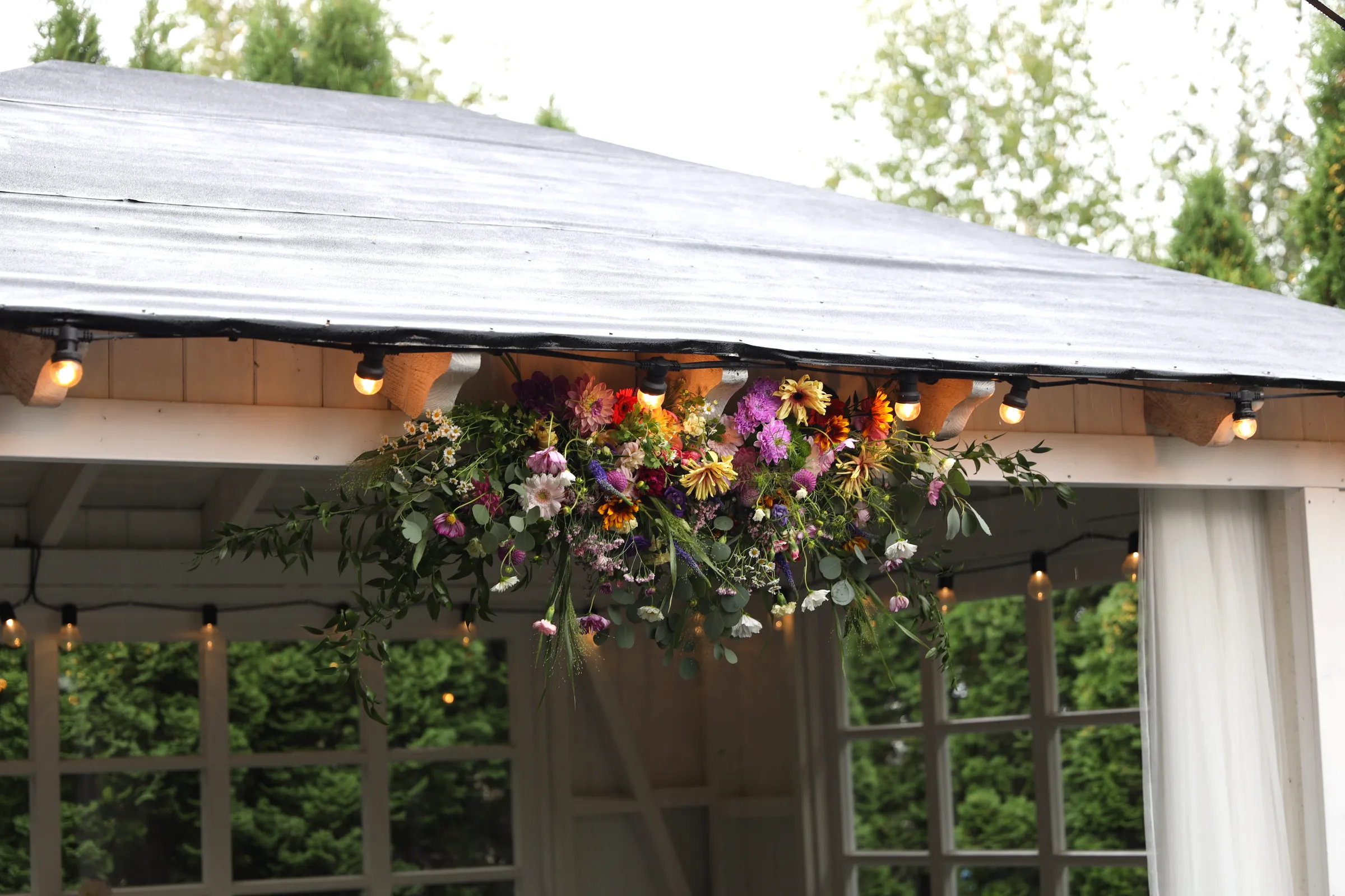 A loose composition of dahlias, cosmoses, gerberas and greenery suspended under the eaves of a white gazebo with a gray roof and lamps