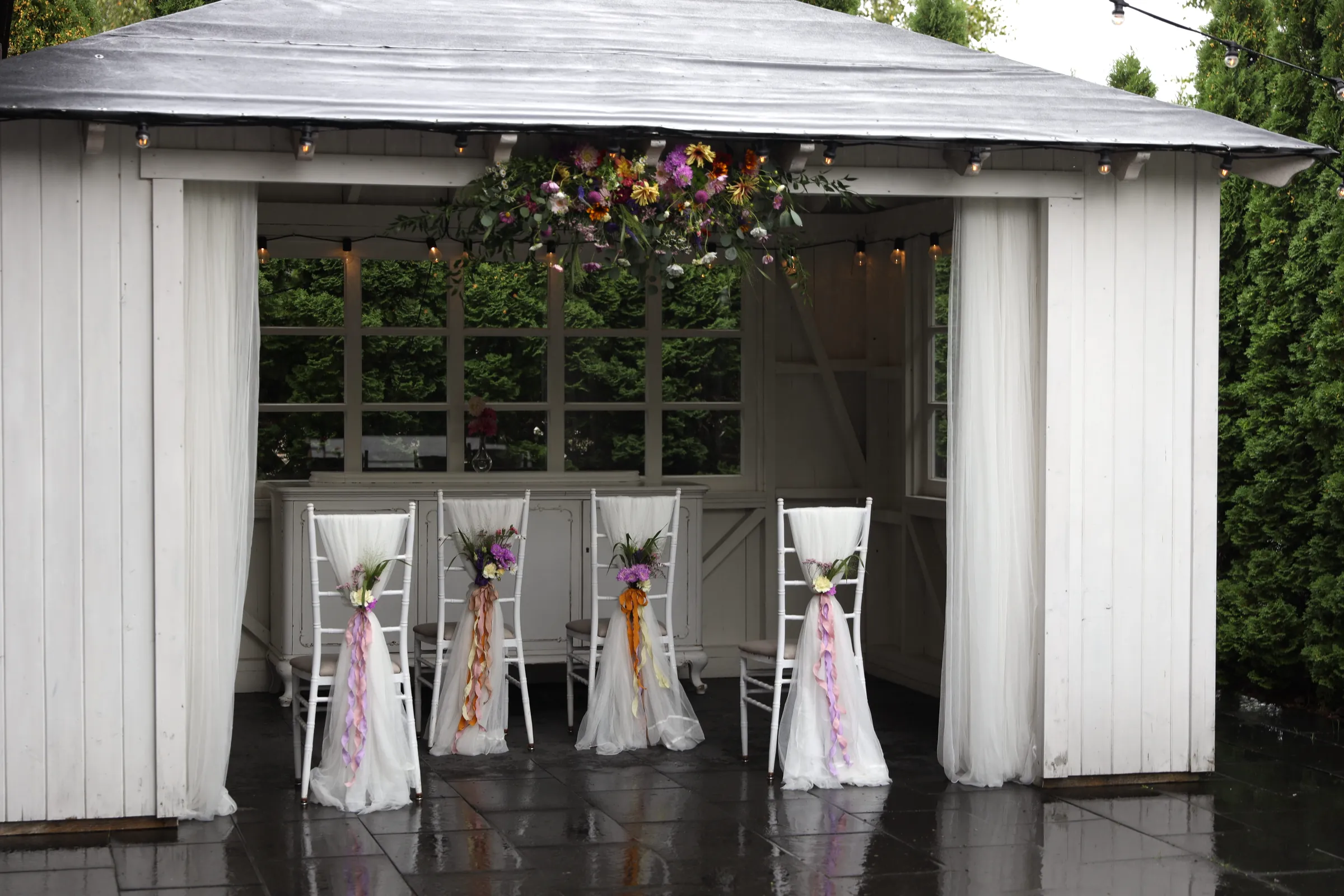 Four white tulle chairs with bouquets and colorful ribbons in the gazebo, above them a composition of wild flowers
