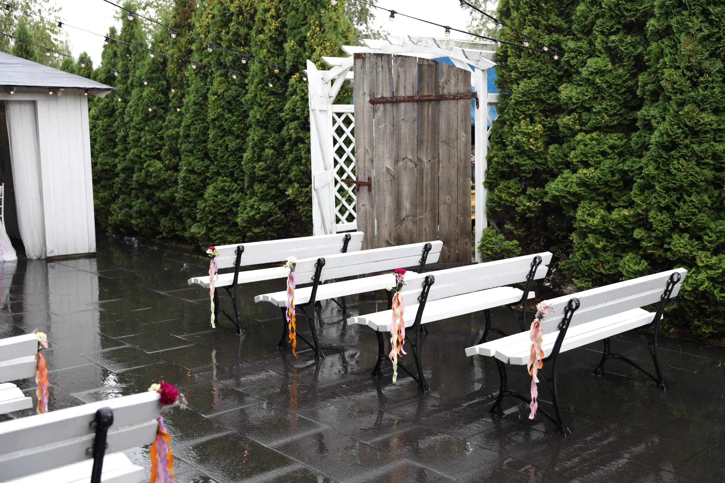 White benches on black cast iron legs with bouquets and long ribbons on a wet square between thuja hedges