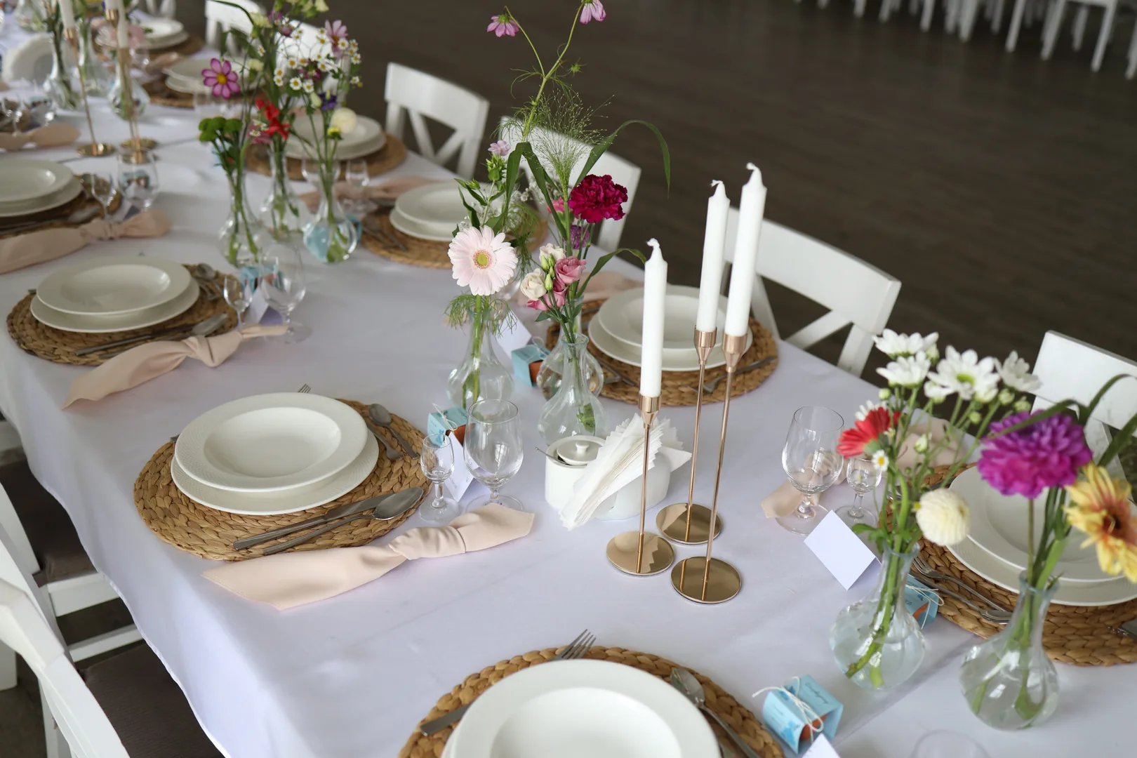 Place setting with a woven placemat and a napkin tied in a knot, next to it are three white candles and vases with a gerbera, a carnation and a cosmos.