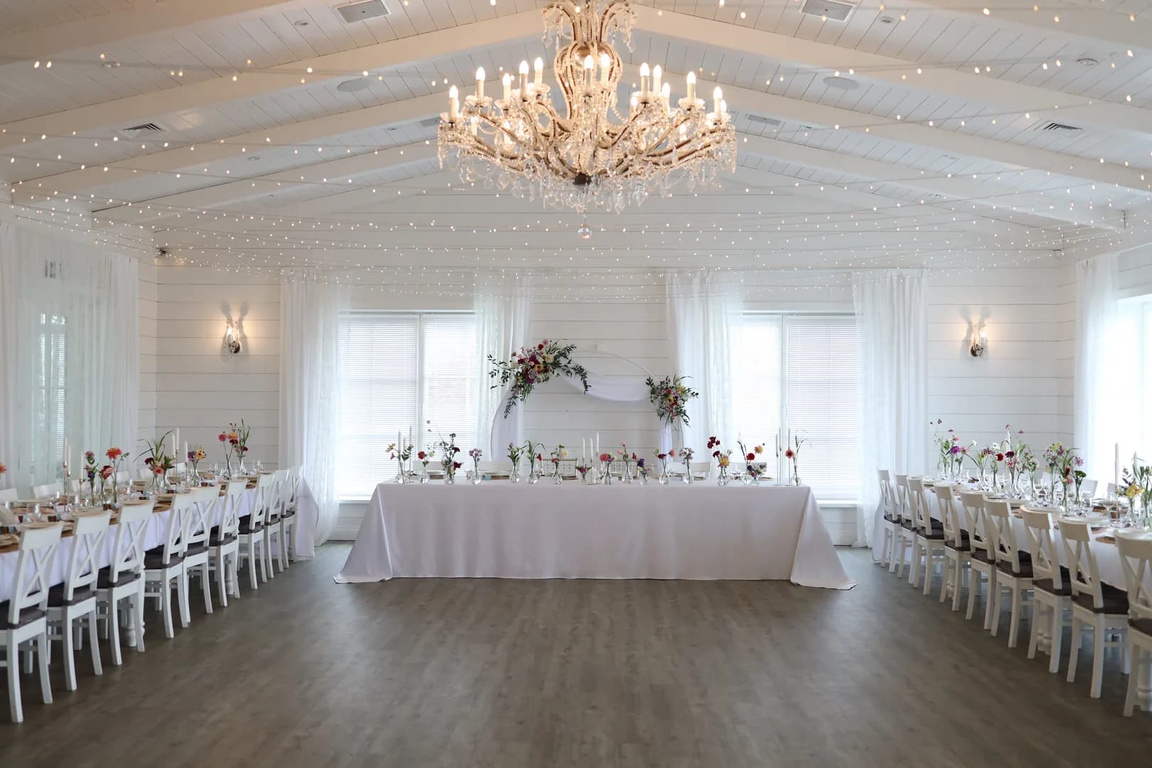 A white room under a crystal chandelier: long tables of guests with wild flowers and a table of the bride and groom in front of a circle with tulle