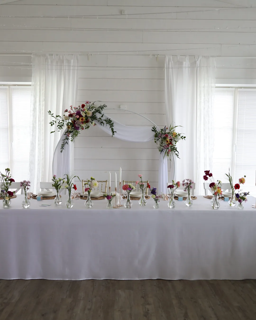 The bride and groom's table with a row of vases with wild flowers and candles, behind it a white circle with tulle and two compositions