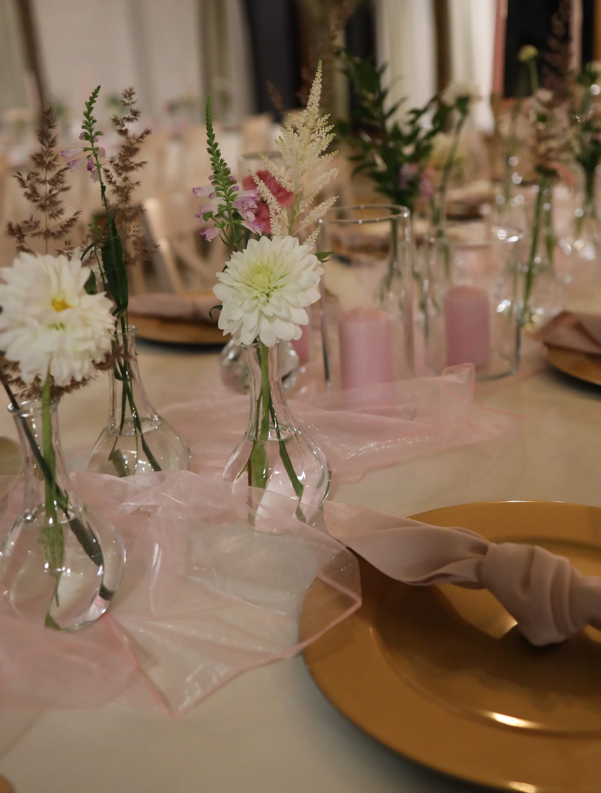 White dahlias in vases, pink flowers on tall shoots and ears of grass, next to them are thick pink candles in glass cylinders and a tulle table runner.