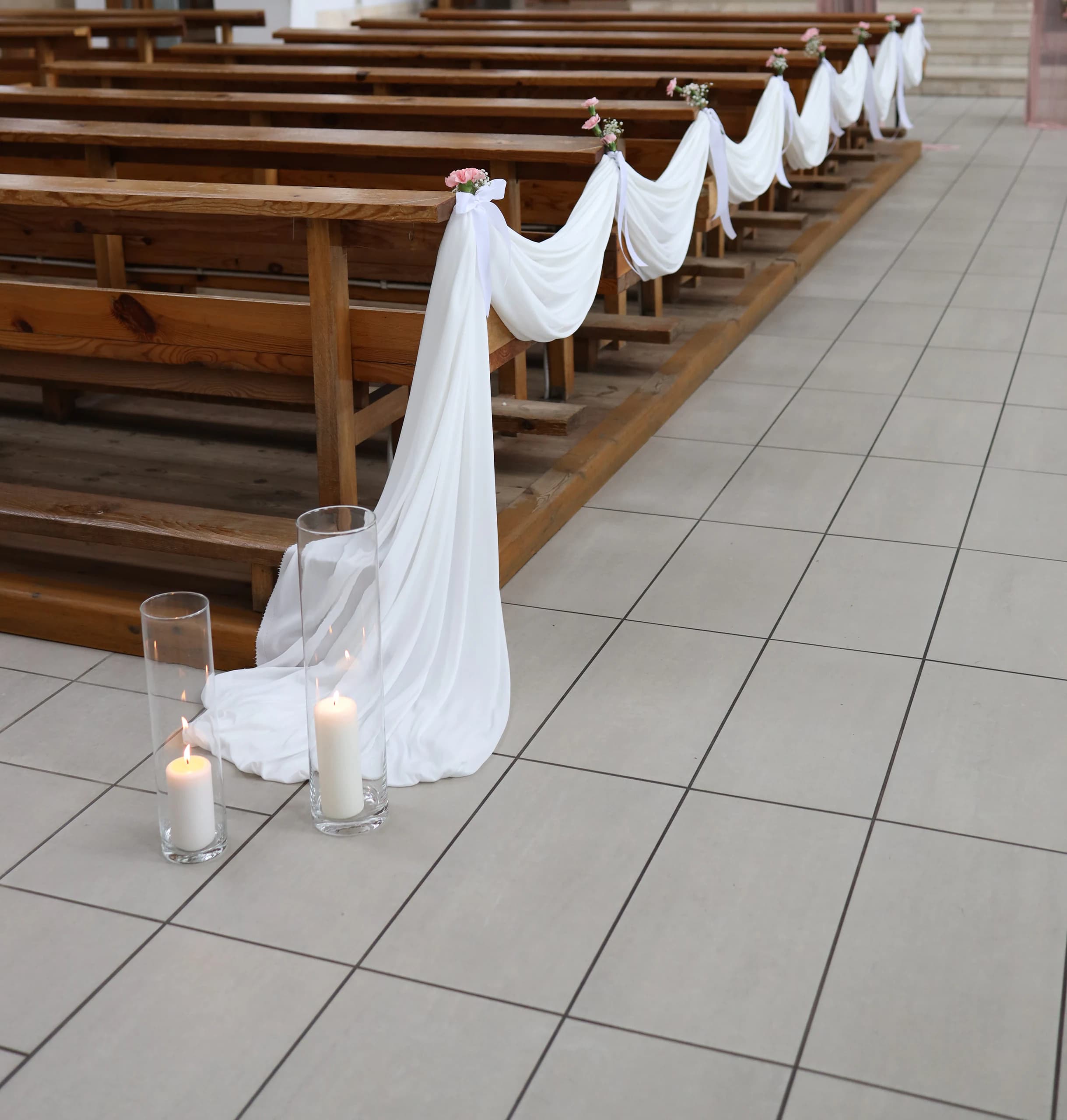 Benches from the side: white fabric flows onto the floor, white bows and pink carnations with gypsophila at the ends of the benches, lit candles on the floor