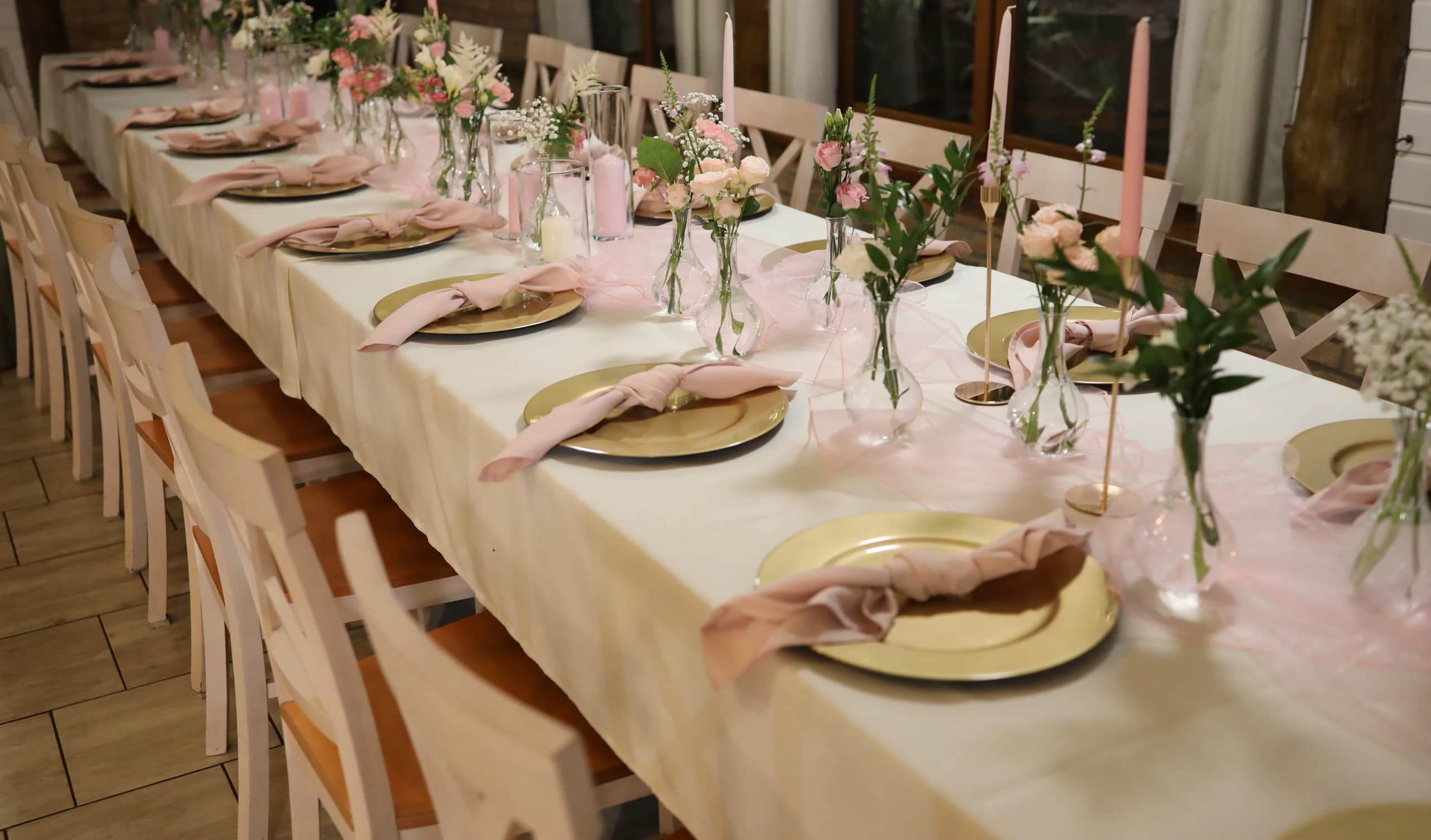 A long table of guests from the side and above: a row of gold plates with knotted napkins, a tulle table runner, vases, pink candles and white chairs