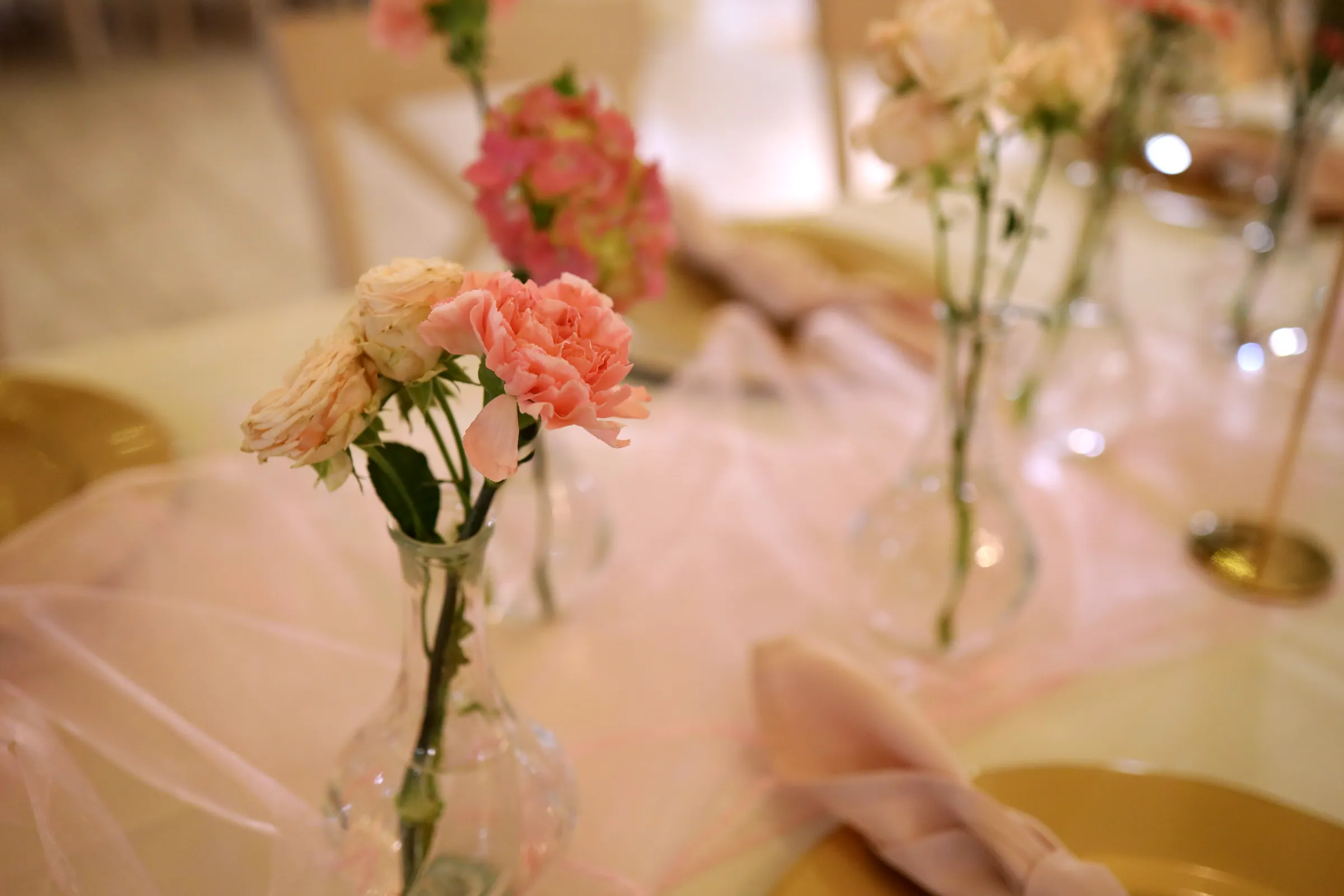 Coral carnation and cream spray roses in a glass vase on pink tulle, pink hydrangea and a gold charger plate in the background
