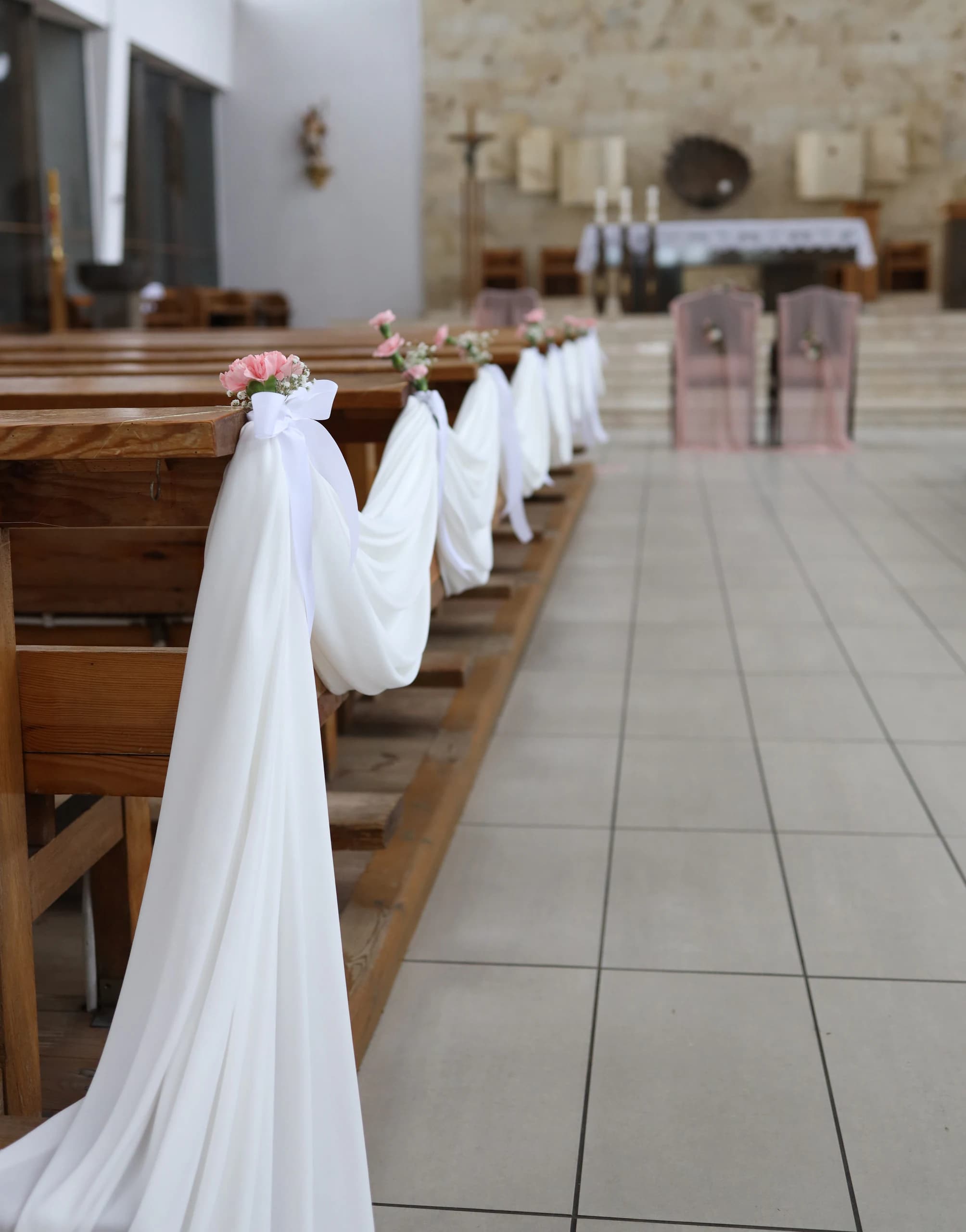 View along the aisle: a cascade of white fabric next to the first bench, bows and pink bouquets on the next ones, chairs and an altar in the background