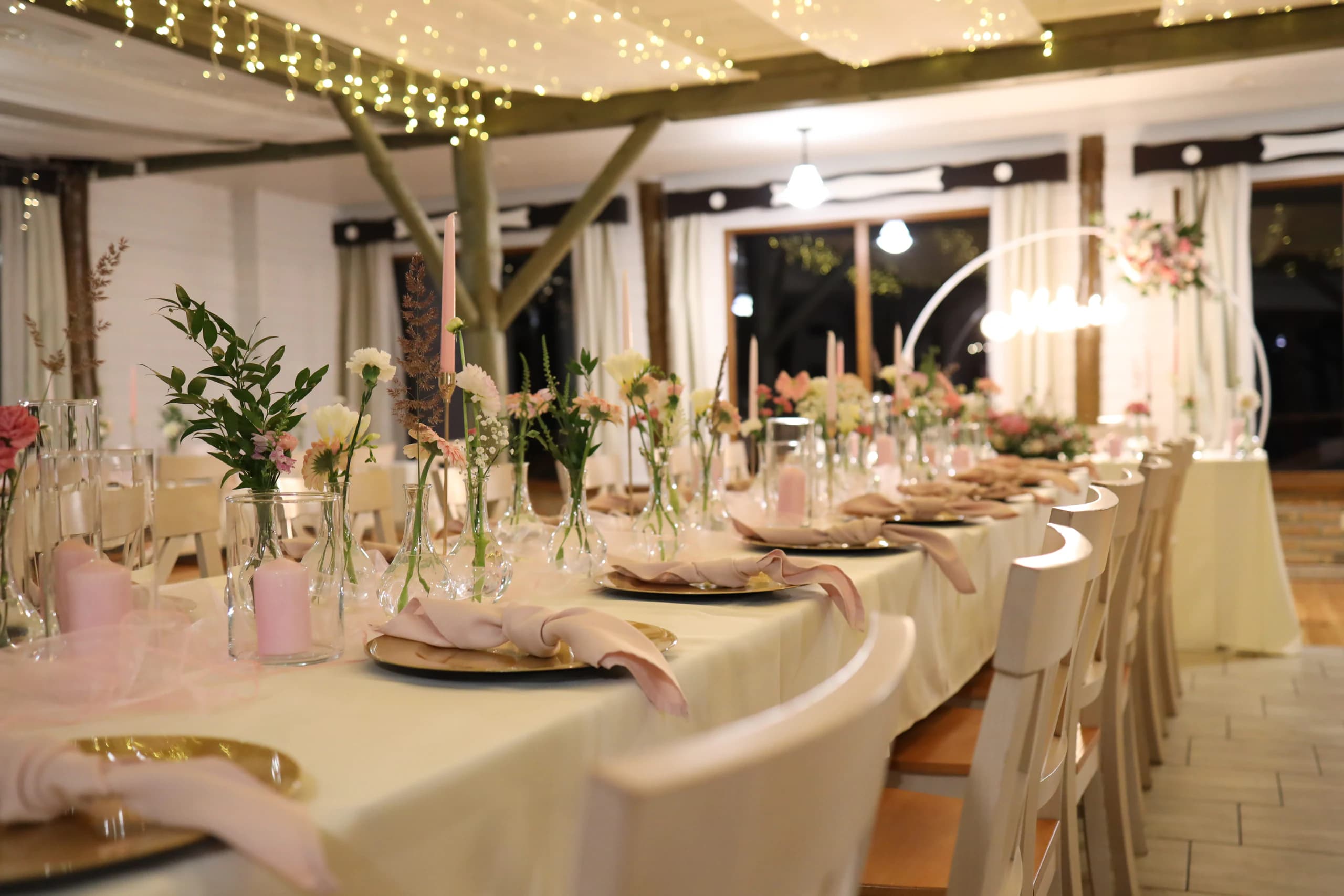 View along the guests' table: vases with carnations, gerberas, twigs and ears of grass, pink candles, in the background a blurred rim with a neon sign