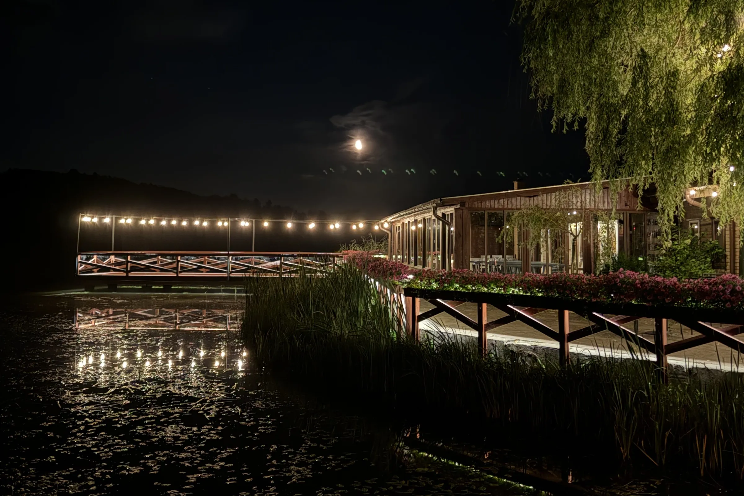 A bridge with a garland of lights over the water next to the wedding hall in Ostrzyce, at night, with the reflection of the lights in the water and the moon in the sky