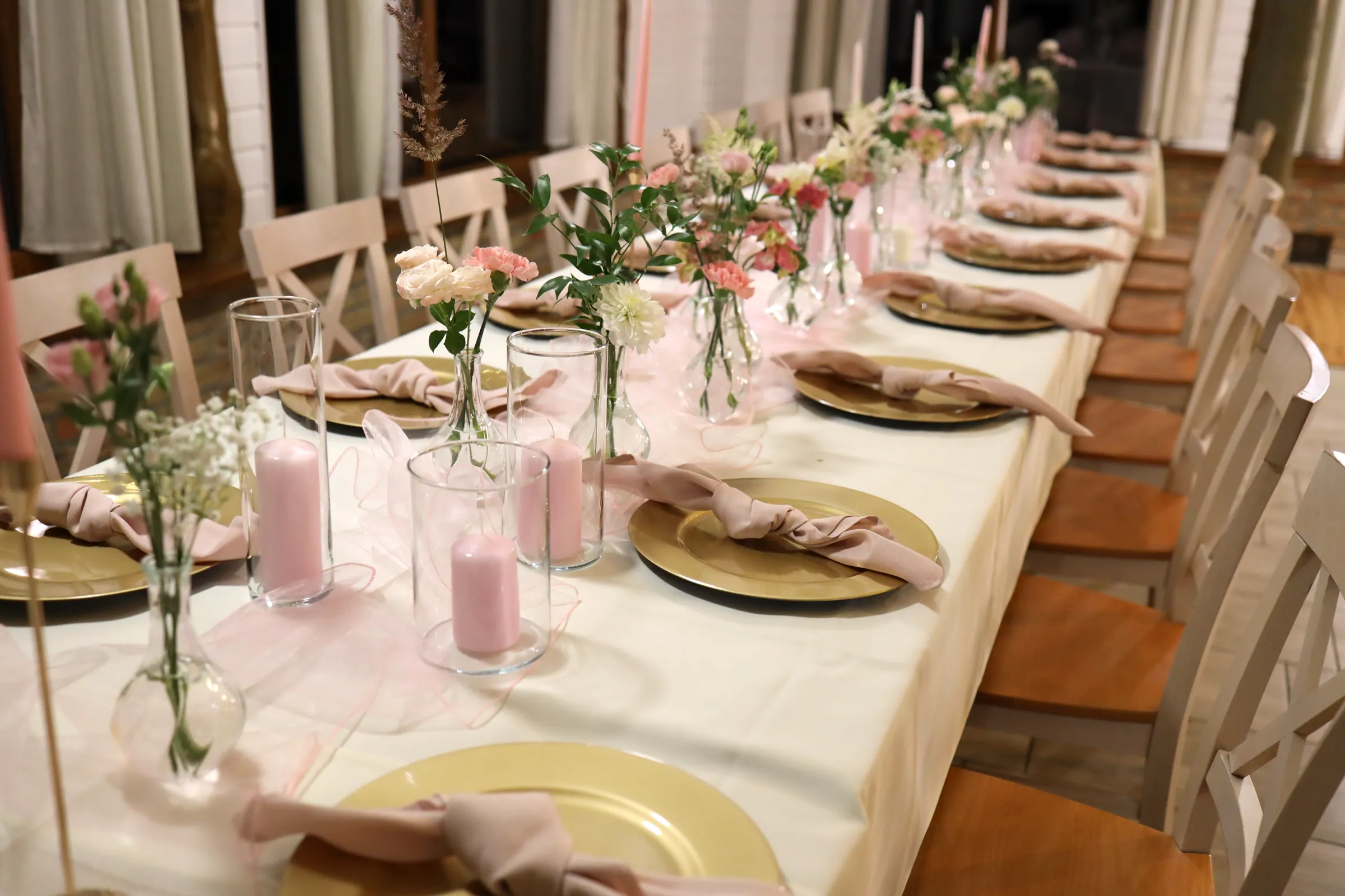 Pink candles in glass cylinders and vases with carnations and gypsophila along a long table with gold charger plates