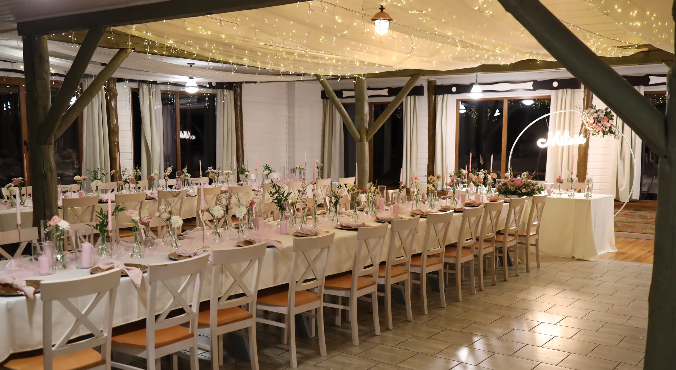 Wide shot of the room: long tables with cream tablecloths, white chairs with cross-backs, on the right the bride and groom's table, lamps from the ceiling
