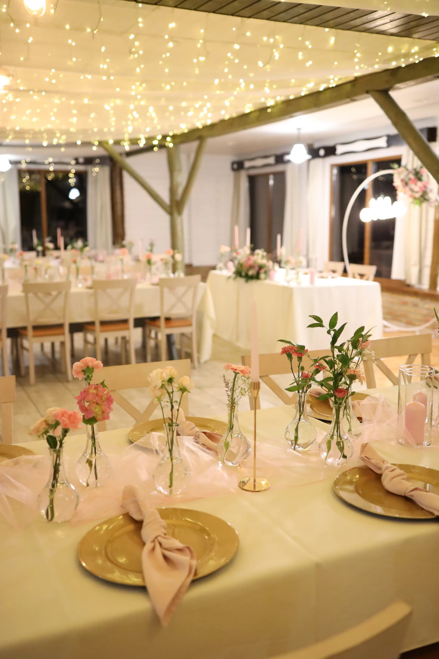 The corner of the guests' table with a gold charger plate, a knotted napkin and vases with carnations, in the background the bride and groom's table with a hoop and a neon sign
