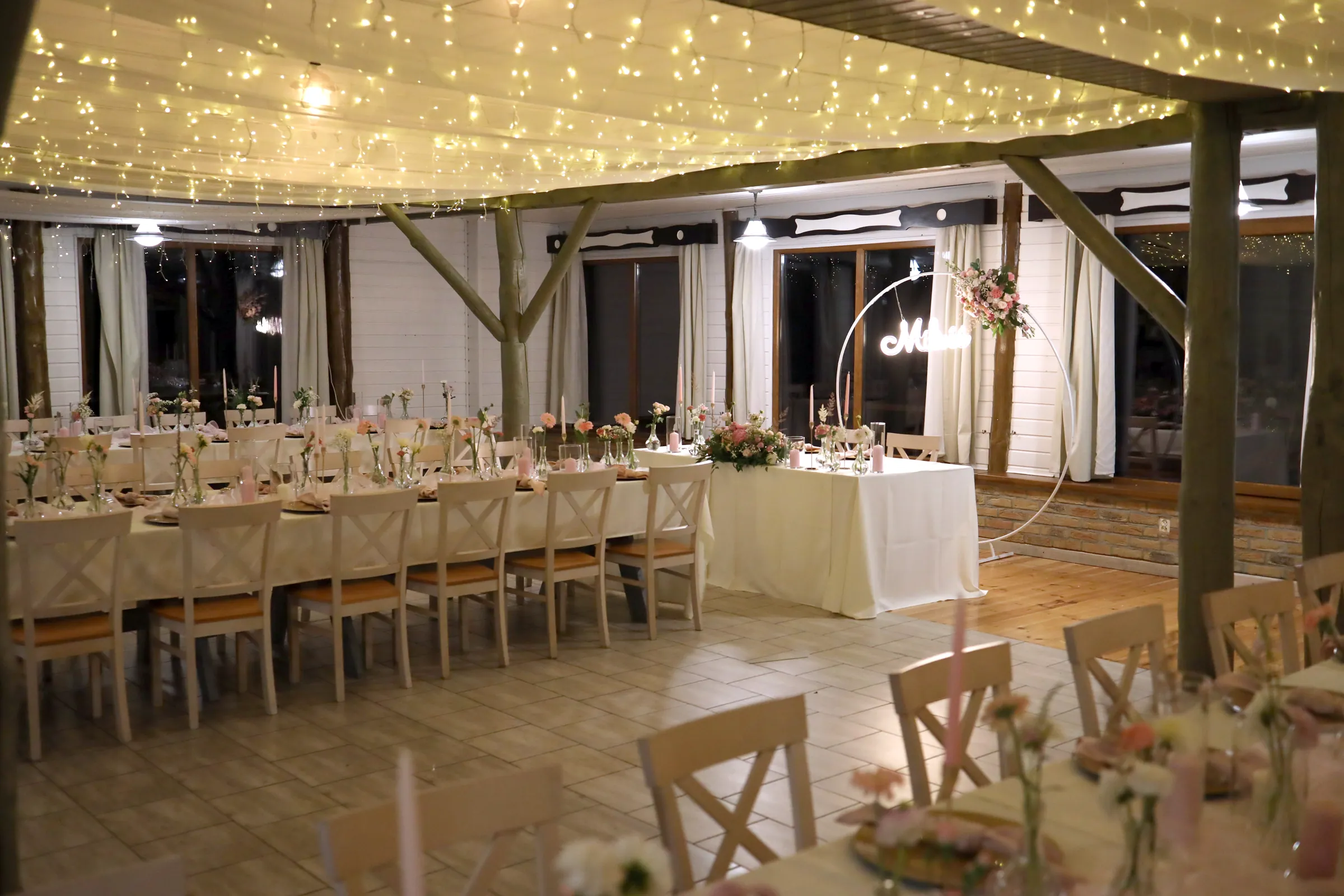 Wedding hall at night under the ceiling with fabric and garlands of lights, long tables of guests and the table of the bride and groom with a white rim and flowers