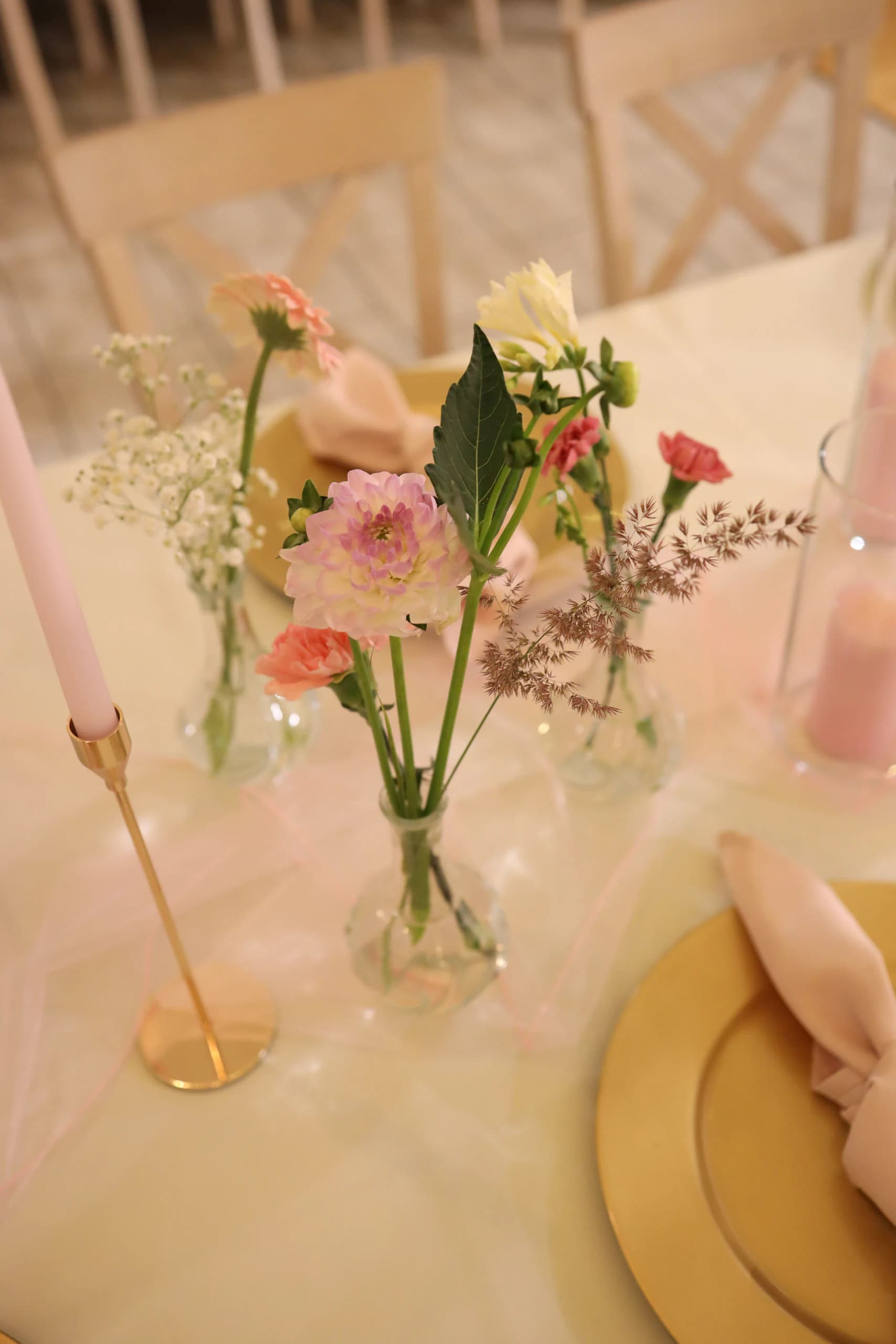 Close-up of vases: pale pink dahlia, coral carnations, cream flower, gypsophila and an ear of grass, next to it a pink candle in a gold candlestick