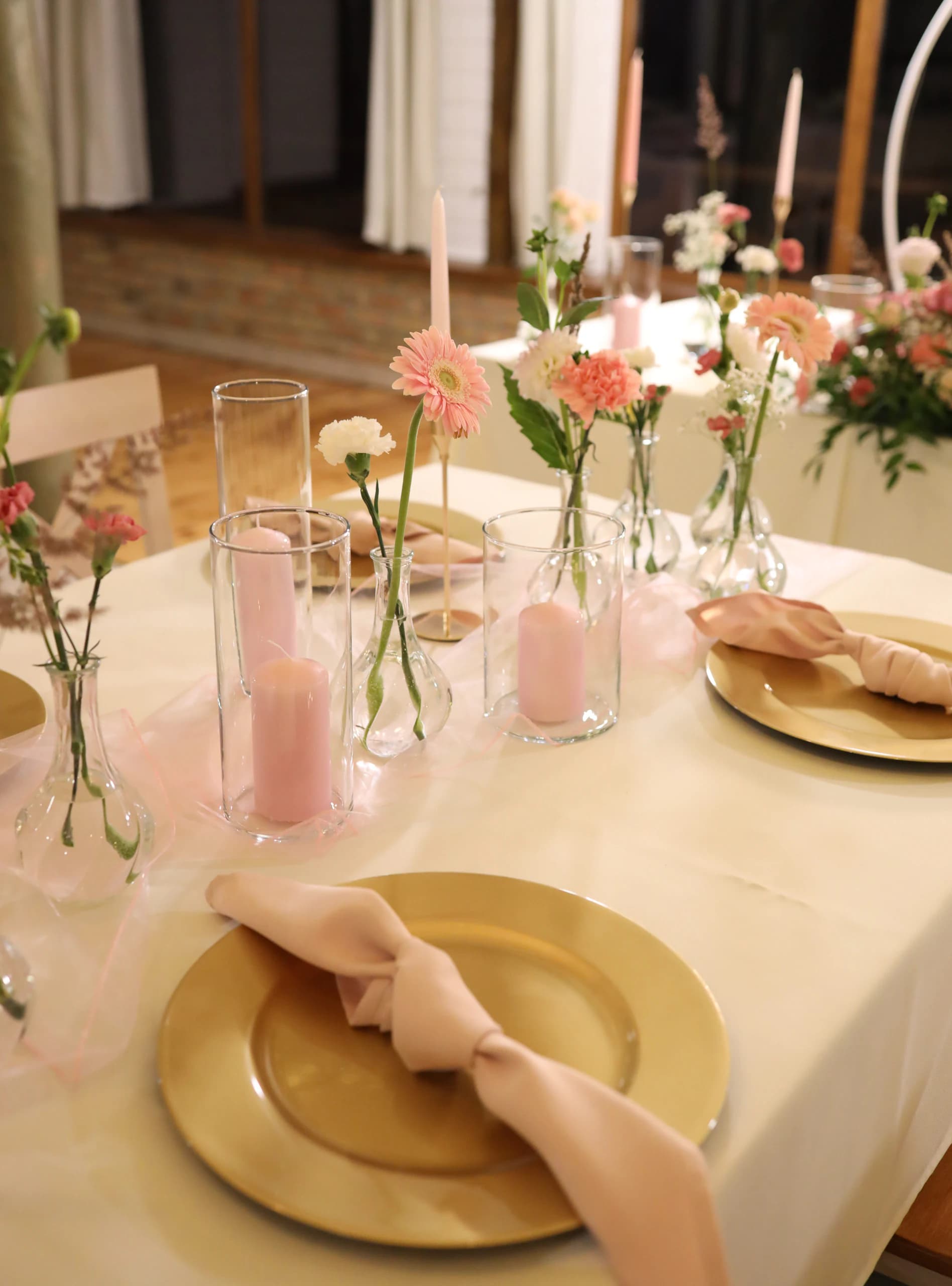 A golden charger plate with a powder pink napkin with a knot, behind it pink candles in glass, vases with gerberas and carnations, in the background the bride and groom's table
