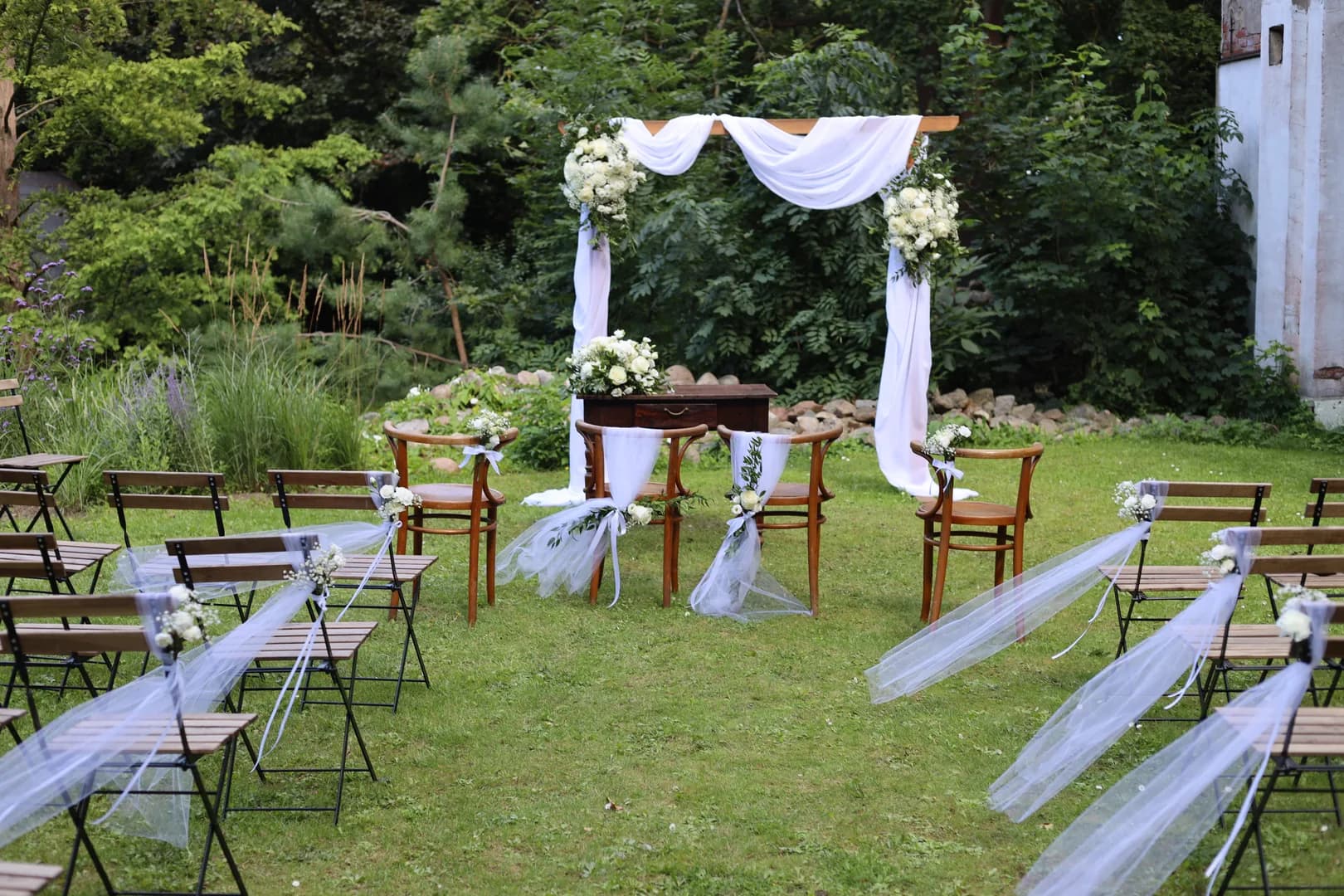 Garden ceremony: wooden arch with white hangings and flowers, officiant's table and tulle chairs by the alley