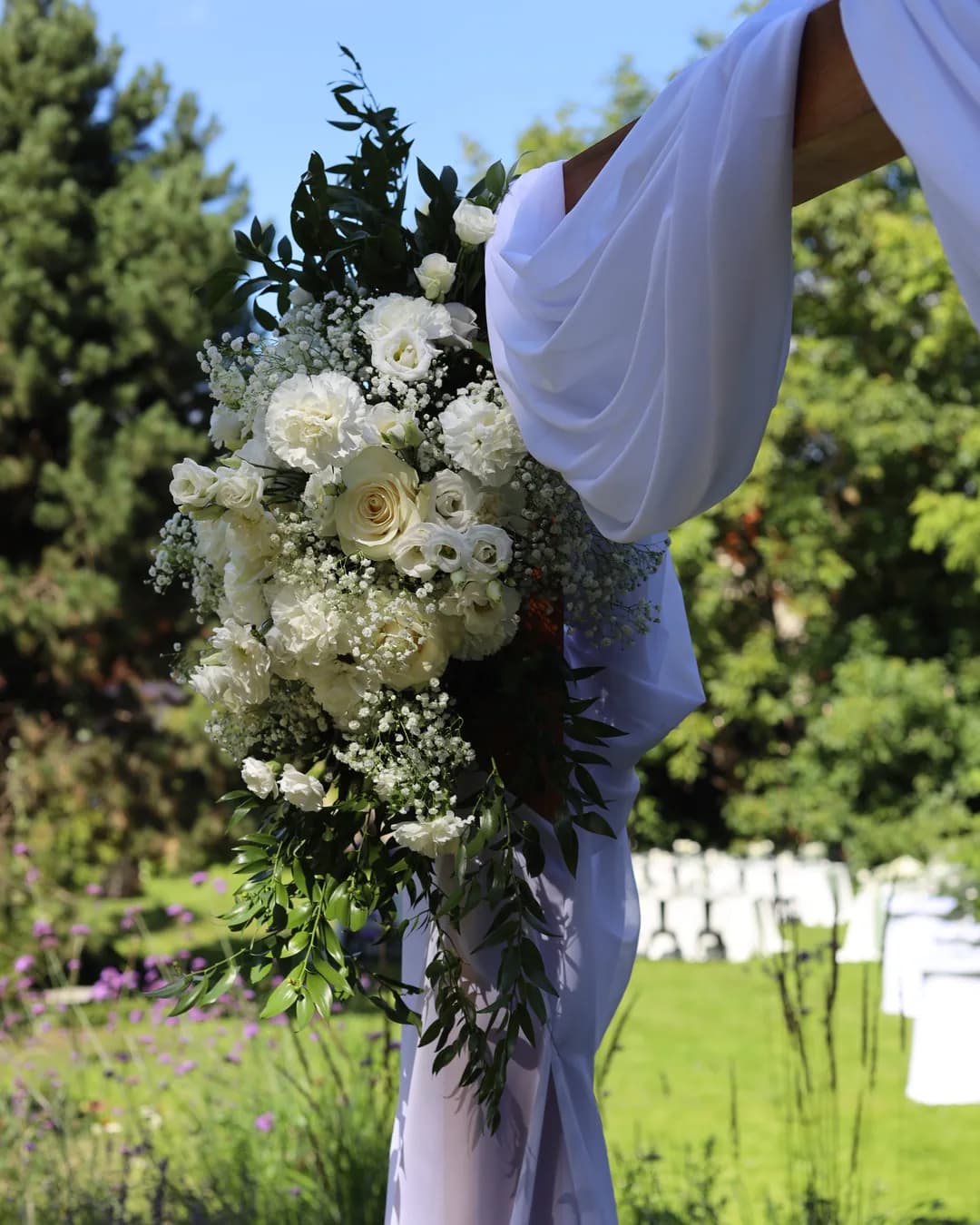 Composition on the corner of an arch of white roses, carnations, eustoma and gypsophila, with a cascade of ruscus against the background of the sky and trees