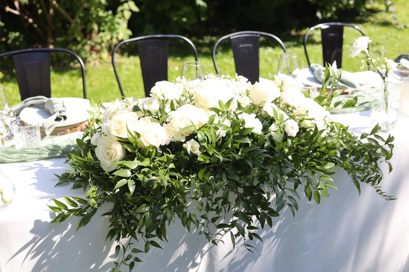 A long, low composition on the bride and groom's table of white roses, eustoma, carnations and gypsophila, with ruscus falling on the tablecloth