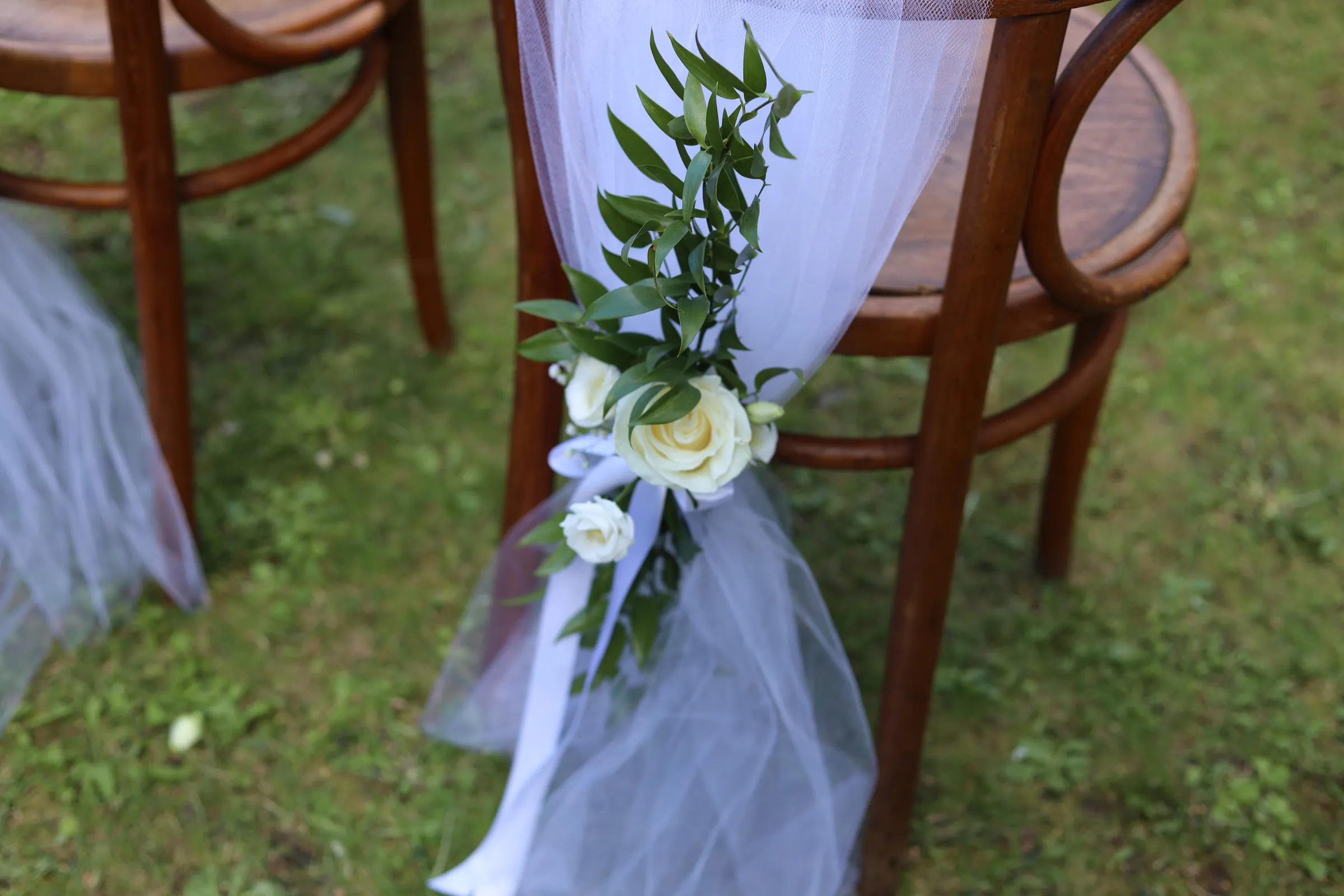 A bent chair wrapped in white tulle, a white rose with smaller flowers, ruscus branches and a white satin ribbon at the backrest.