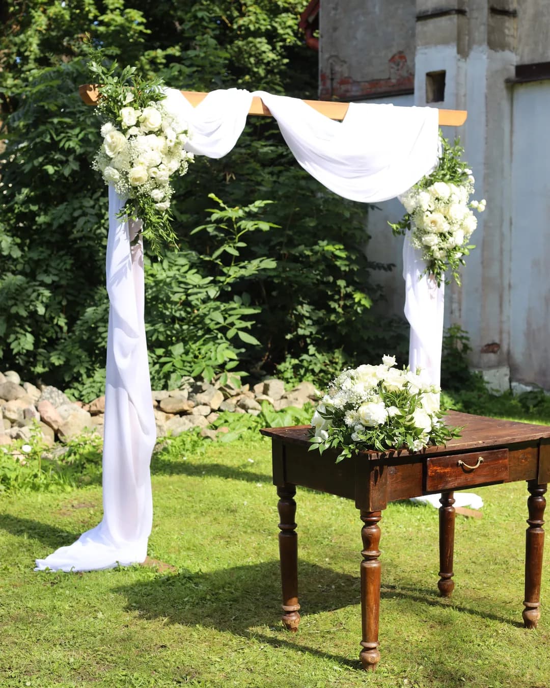 A wooden arch with white draperies and two white flower arrangements, and an official's table with low decoration
