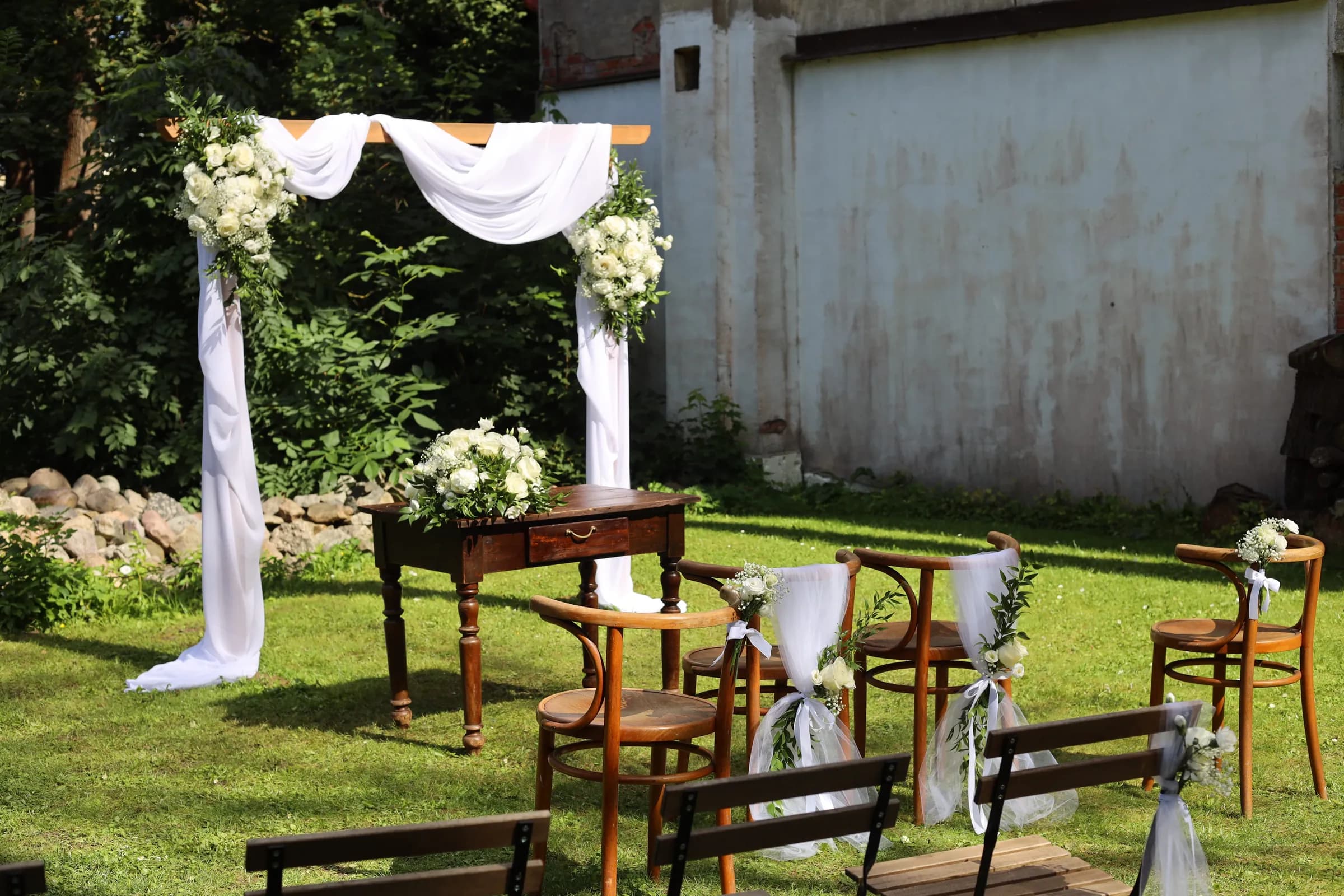 A wooden arch with white drapery and flowers, an official's table with a low composition and bent chairs with tulle near a whitewashed wall